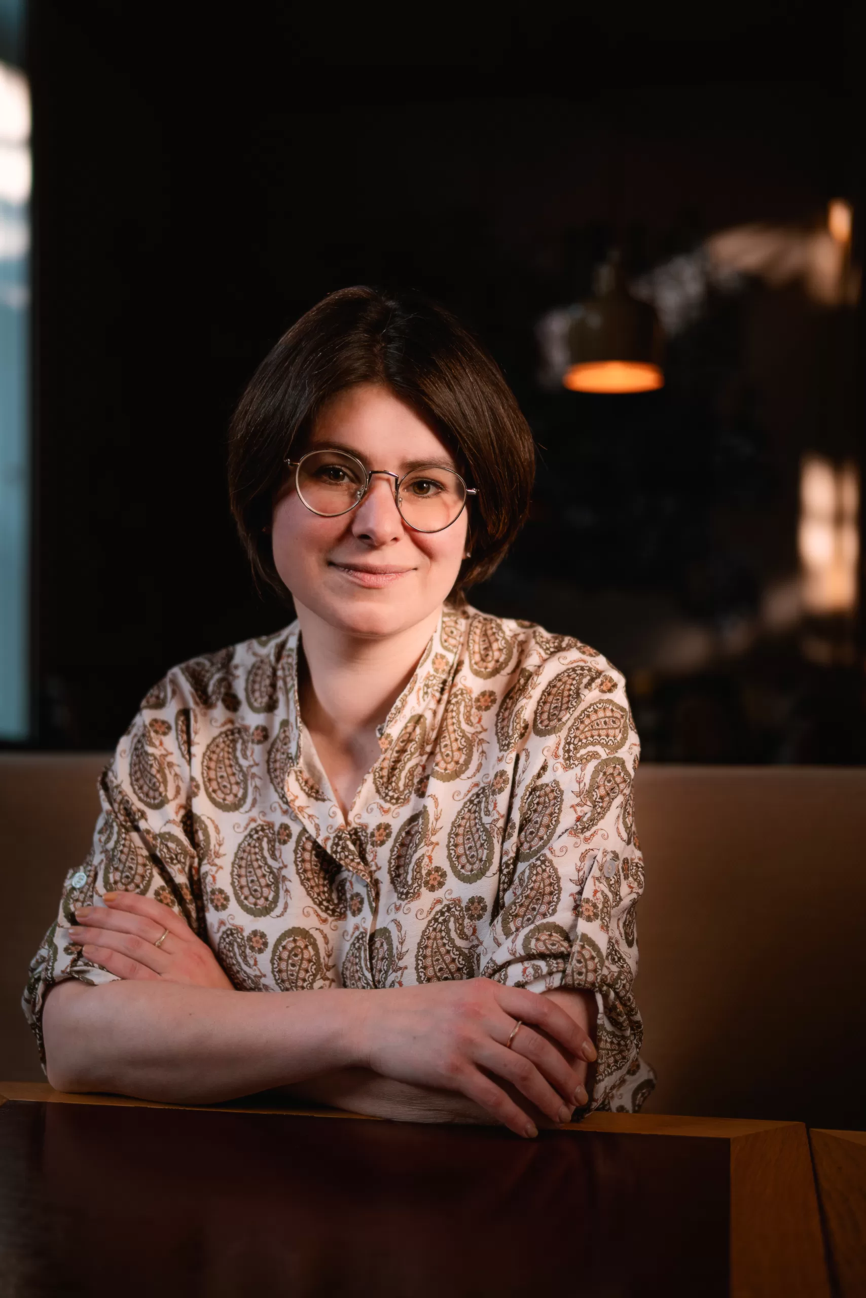 Young woman with short brown hair and glasses wearing a paisley shirt sits at a dark table in a dim, cozy setting.