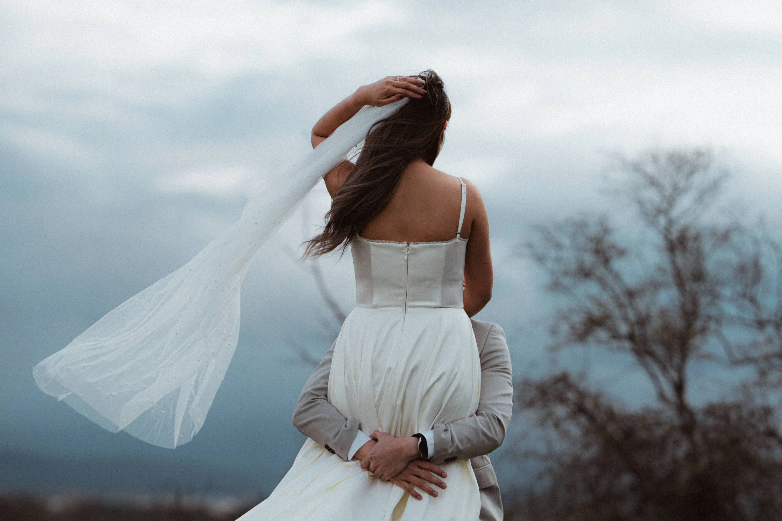 A photo of a bride and a groom holding her in hands on a hill with overcast sky in the background
