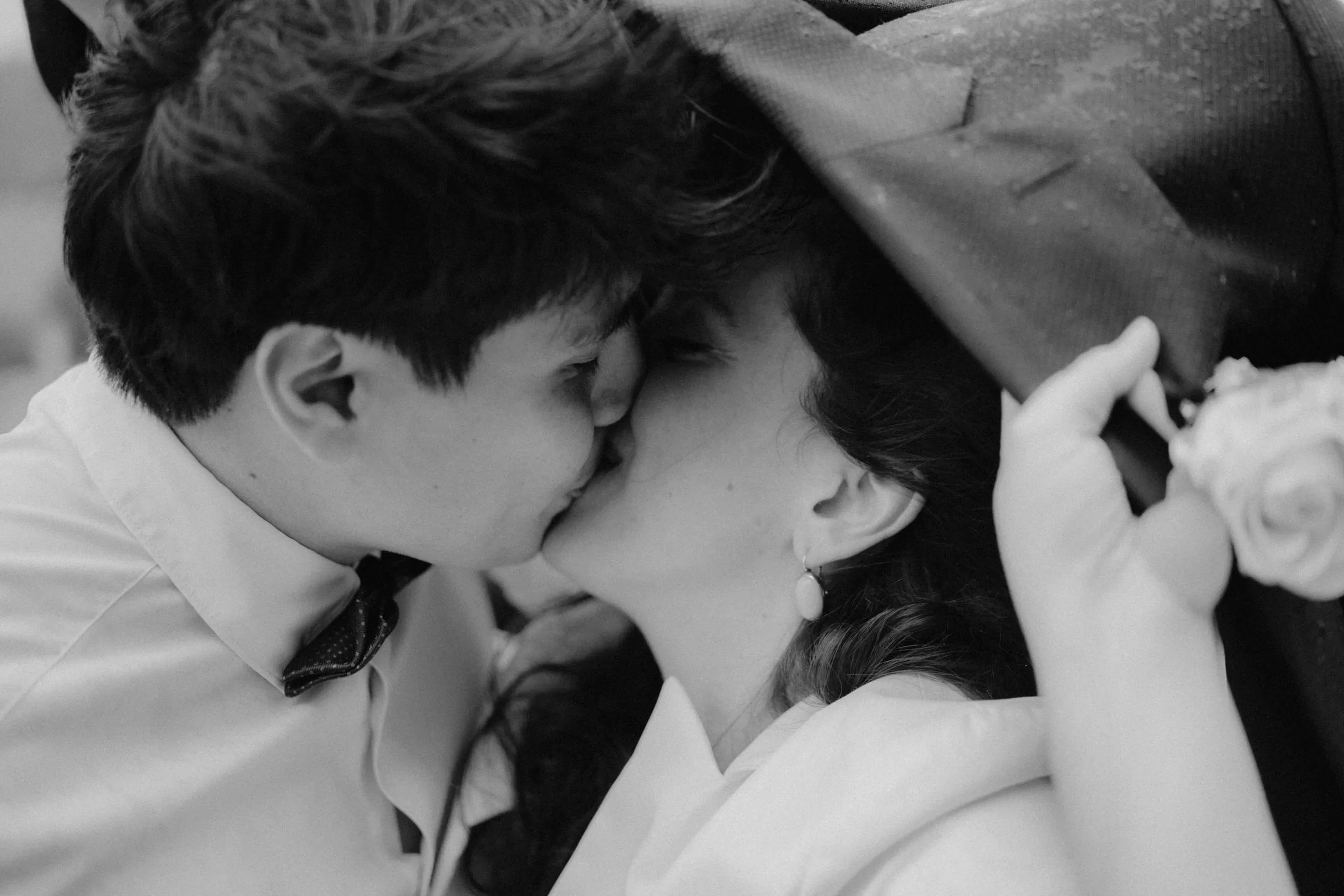 Black and white photo of a couple hiding under a jacket and kissing on a rainy day on their wedding