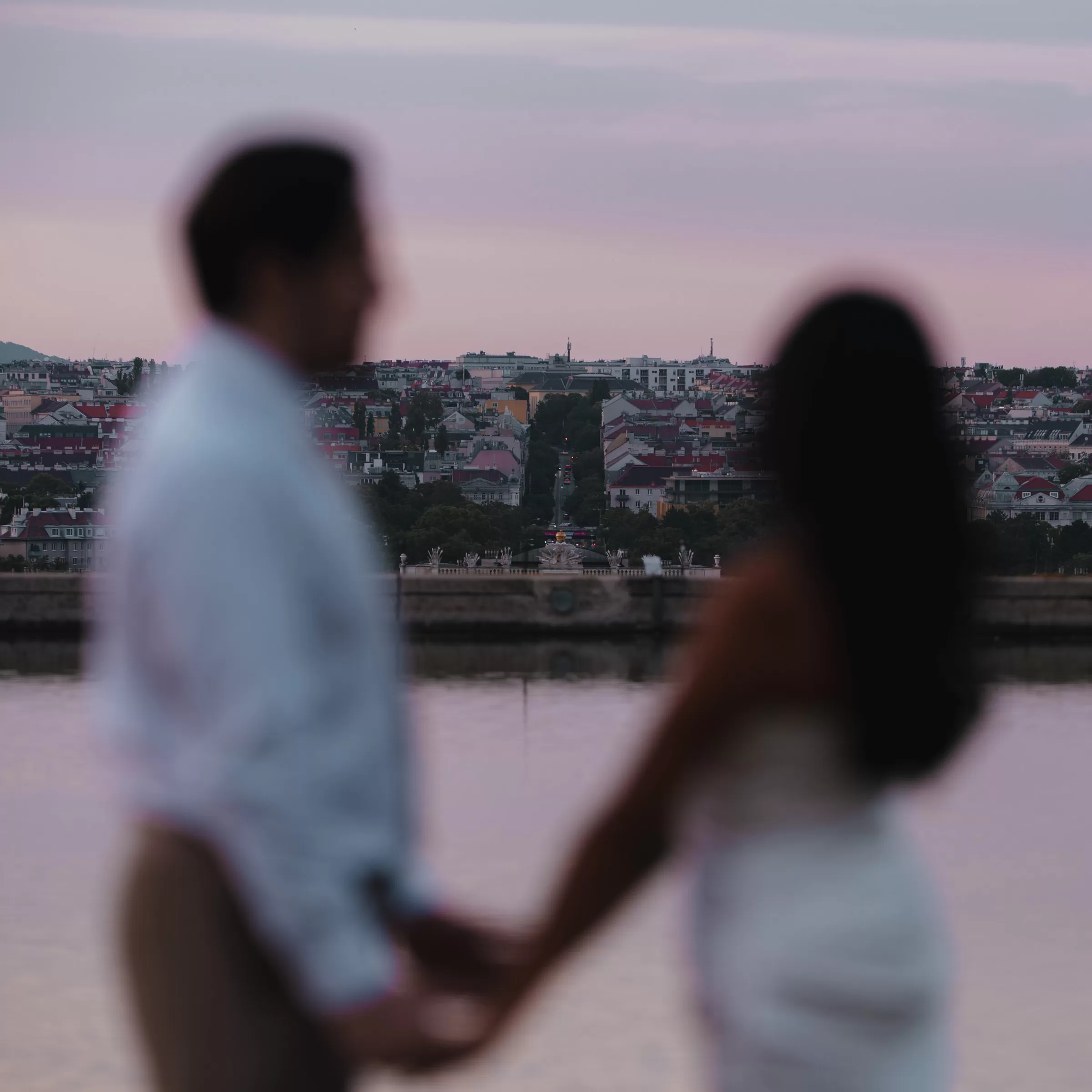Photographer in Berlin Fedor Vasilev Photographer In Vienna out of focus couple standing holding hands in front of a pond and a view over the city at sunset