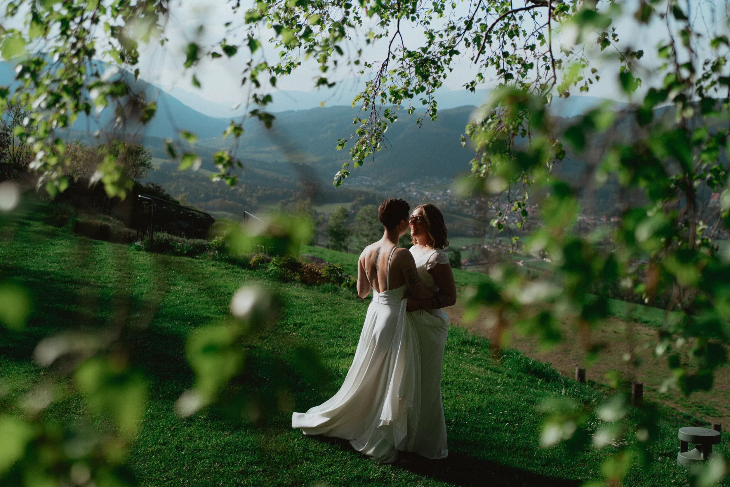 Brides in white dresses dance on a grassy hill in front of Fernblick hotel with the view over the mountains