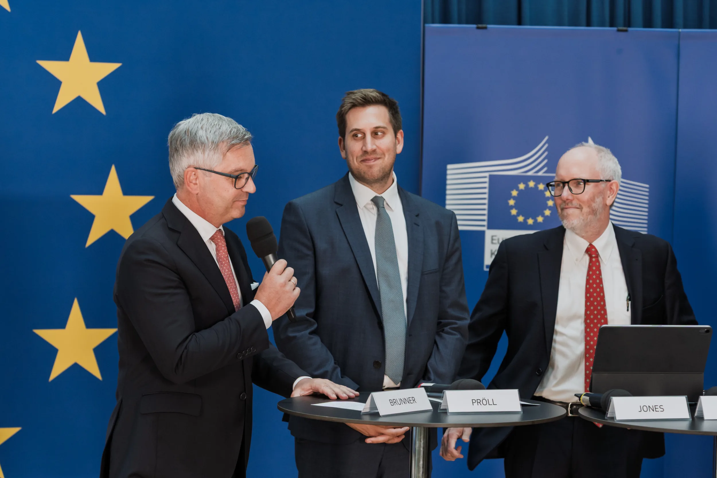 European Union Event Photographer | Grant Signing Ceremony in Vienna Fedor Vasilev Photographer In Berlin Man speaks into a microphone at a table with nameplates for Brunner, Proll, and Jones during an EU grant ceremony.