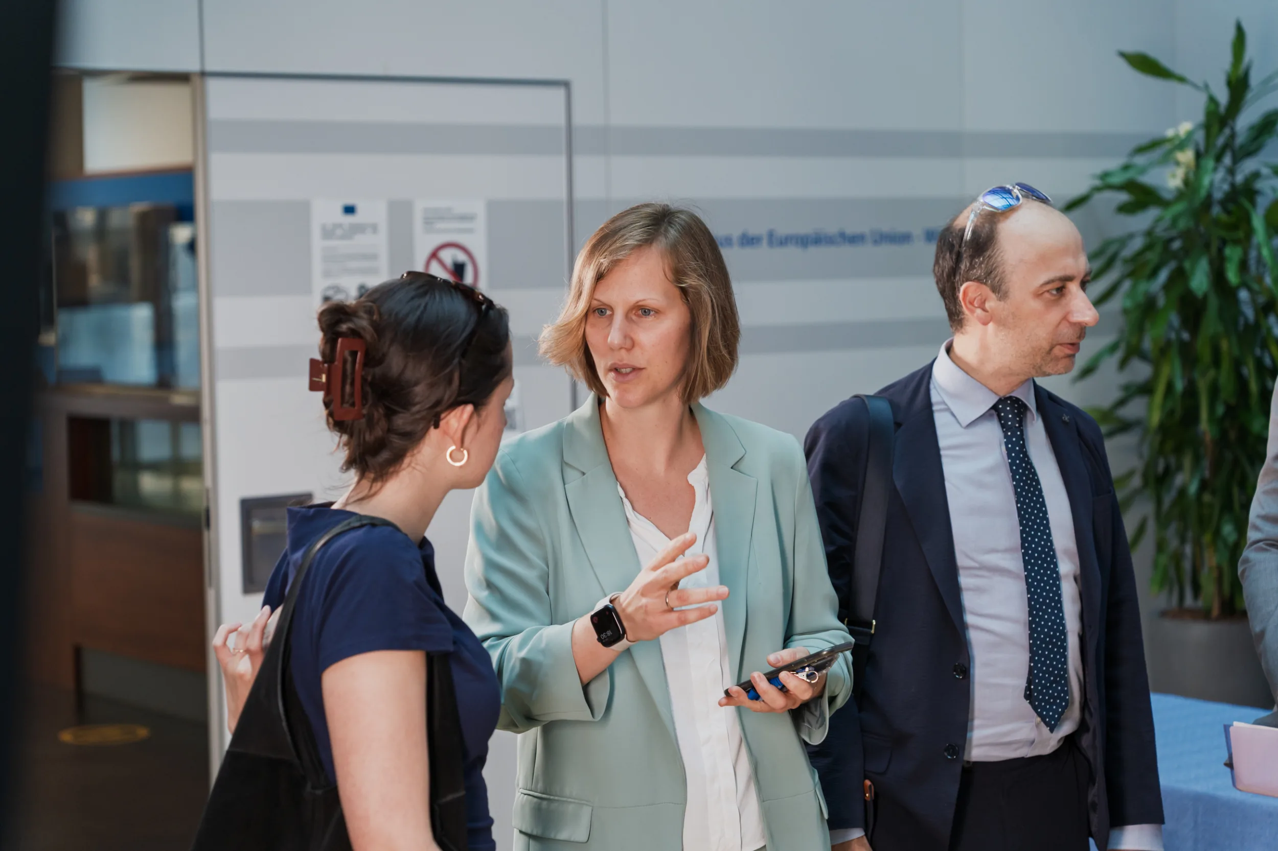 European Union Event Photographer | Grant Signing Ceremony in Vienna Fedor Vasilev Photographer In Berlin Three people in business attire conversing in a bright office lobby at a European Union event.