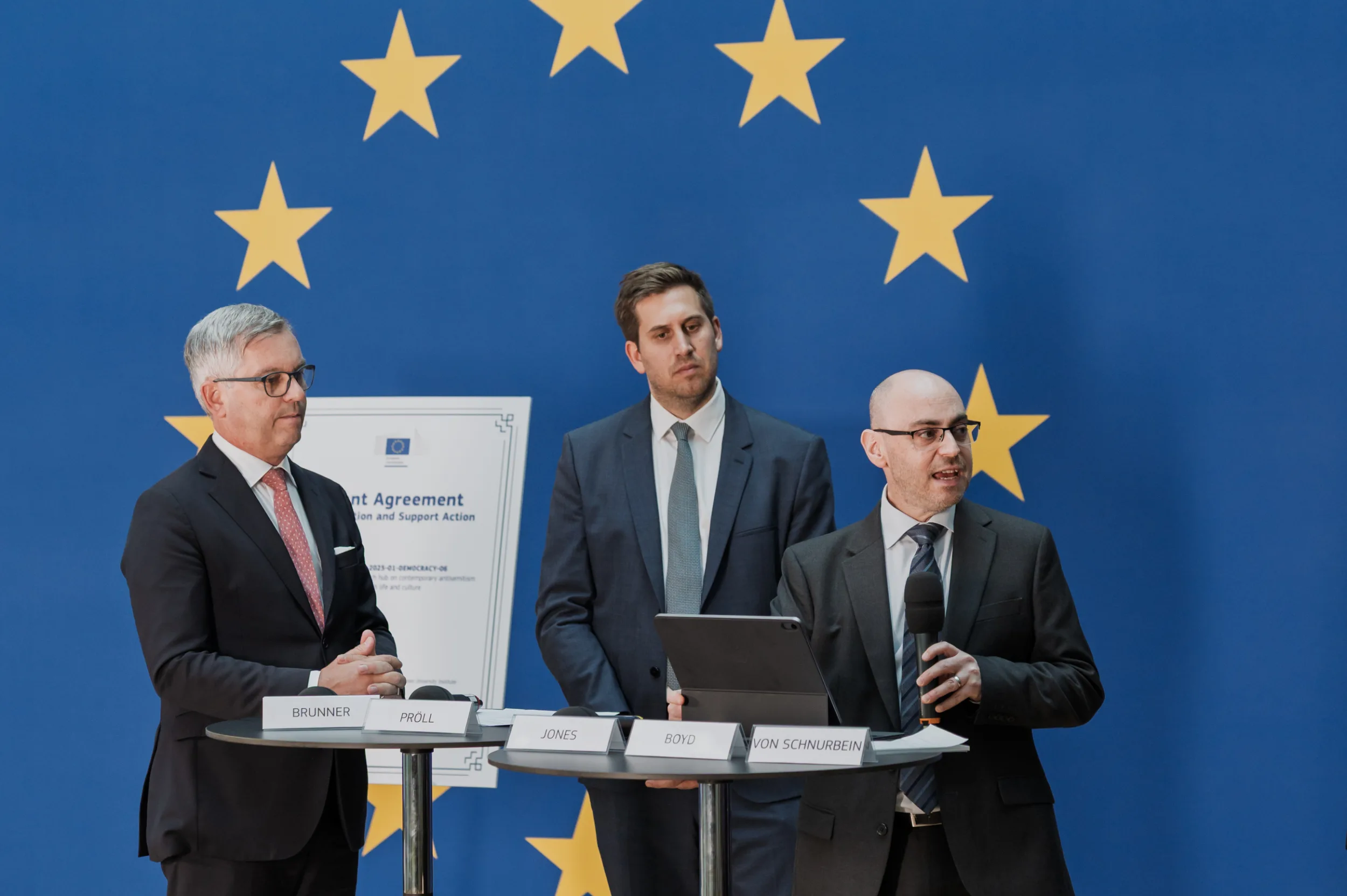 European Union Event Photographer | Grant Signing Ceremony in Vienna Fedor Vasilev Photographer In Berlin Three men stand behind tables labeled with names before a blue European Union background with gold stars.
