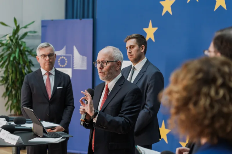 An EU official gestures while holding a microphone at a podium before a blue backdrop with yellow European Union stars.