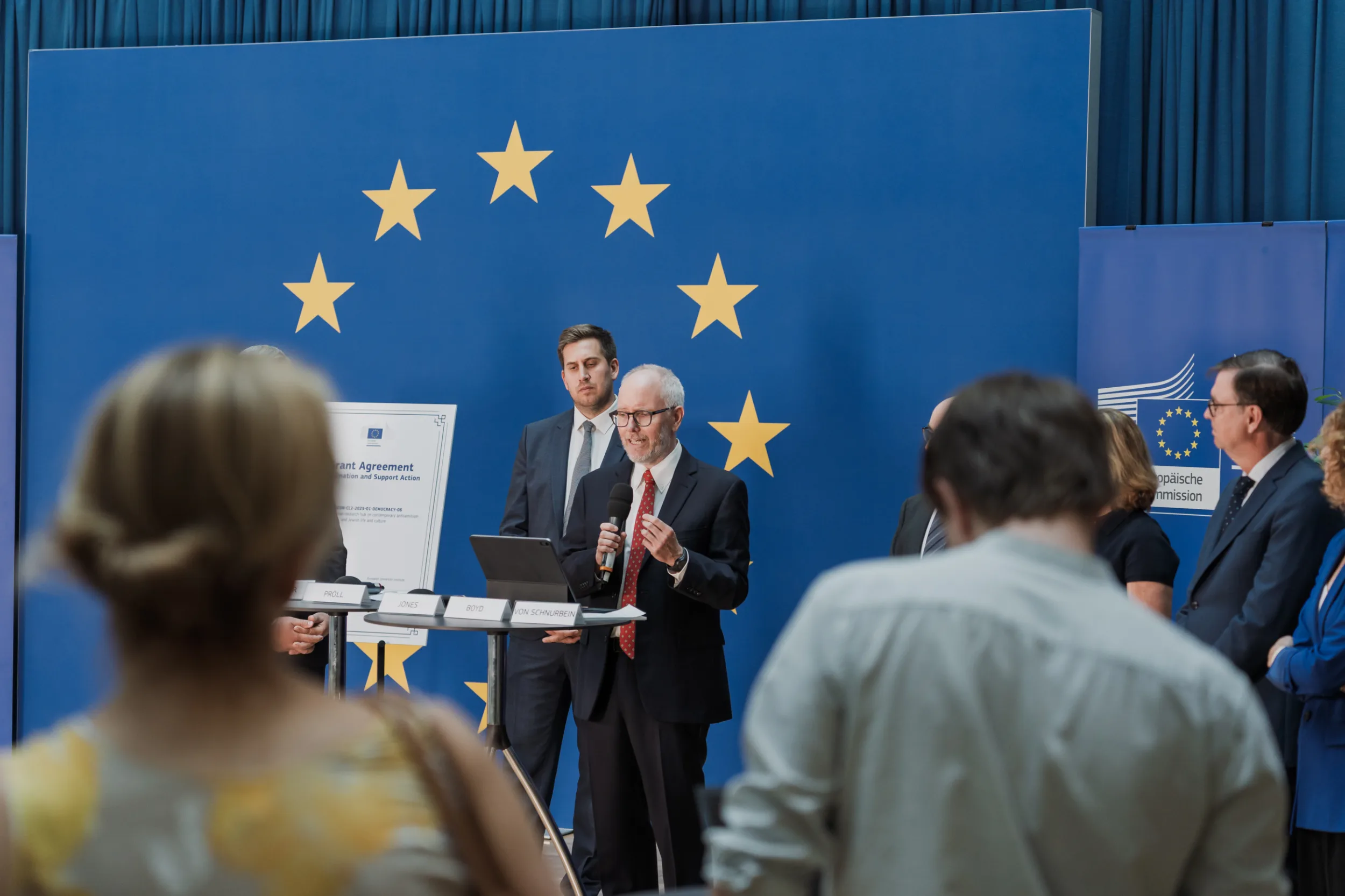 European Union Event Photographer | Grant Signing Ceremony in Vienna Fedor Vasilev Photographer In Berlin A politician gestures while speaking into a microphone before a blue backdrop displaying European Union stars.