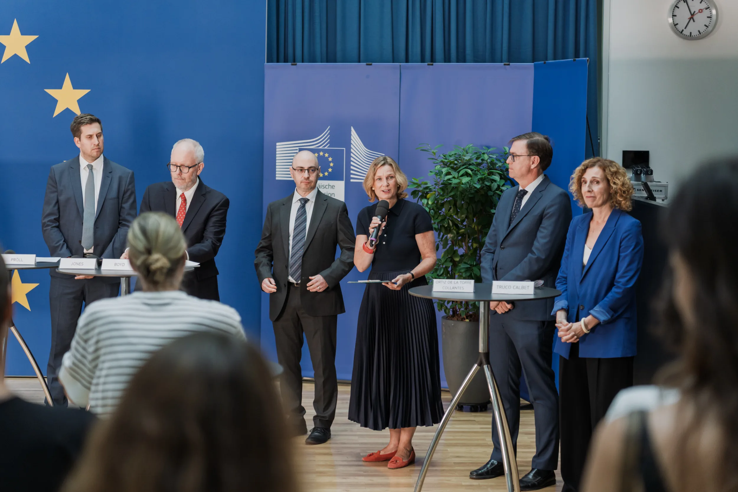 European Union Event Photographer | Grant Signing Ceremony in Vienna Fedor Vasilev Photographer In Berlin Woman in a dark dress speaks into a microphone before a group of professionals standing by European Union backdrops.
