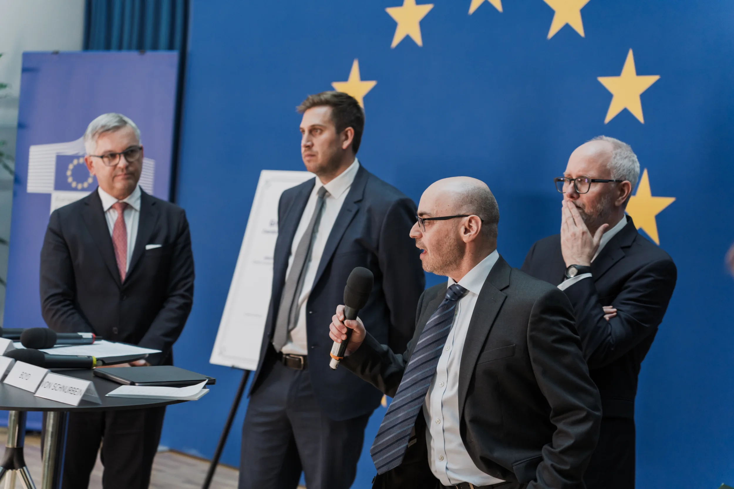 European Union Event Photographer | Grant Signing Ceremony in Vienna Fedor Vasilev Photographer In Berlin An EU representative in a dark suit speaks into a microphone while three colleagues listen during a grant signing ceremony