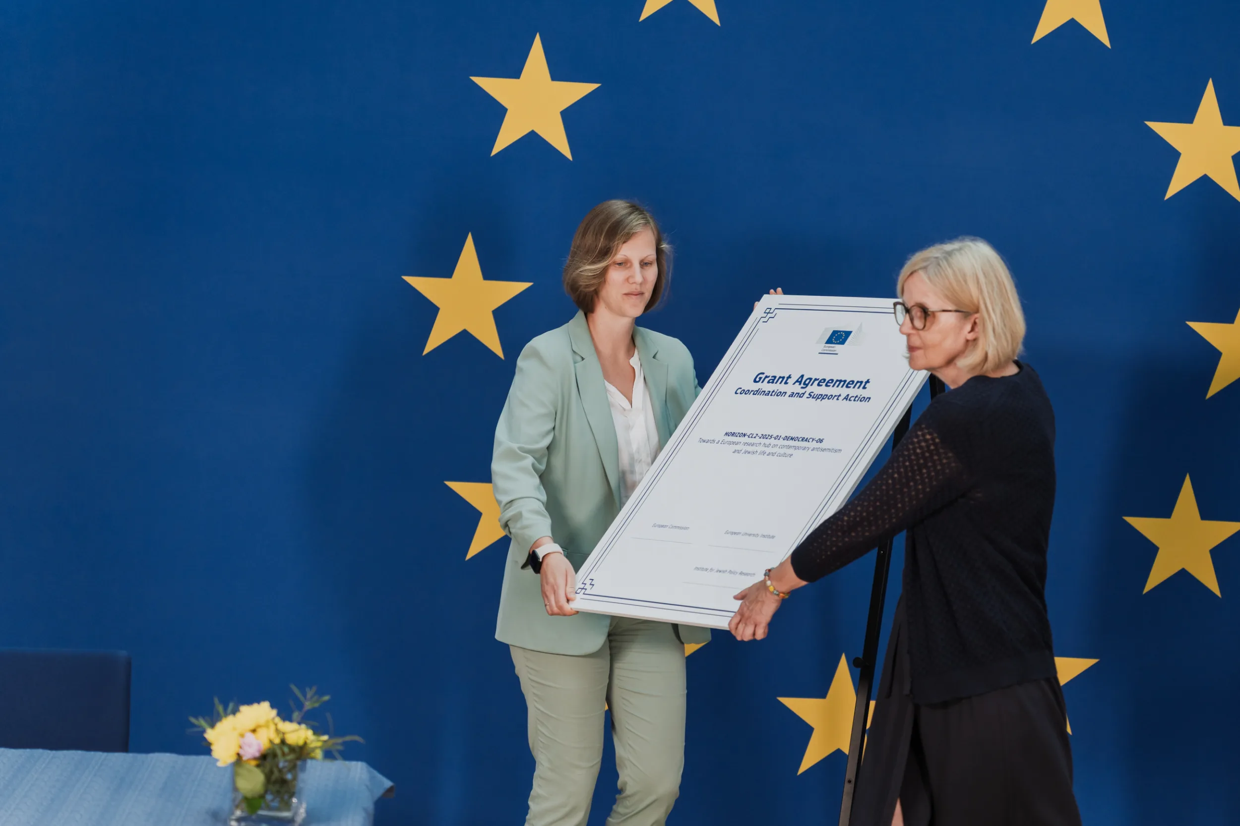 European Union Event Photographer | Grant Signing Ceremony in Vienna Fedor Vasilev Photographer In Berlin Two women adjust a large board displaying a Grant Agreement document before a blue European Union star-themed backdrop.