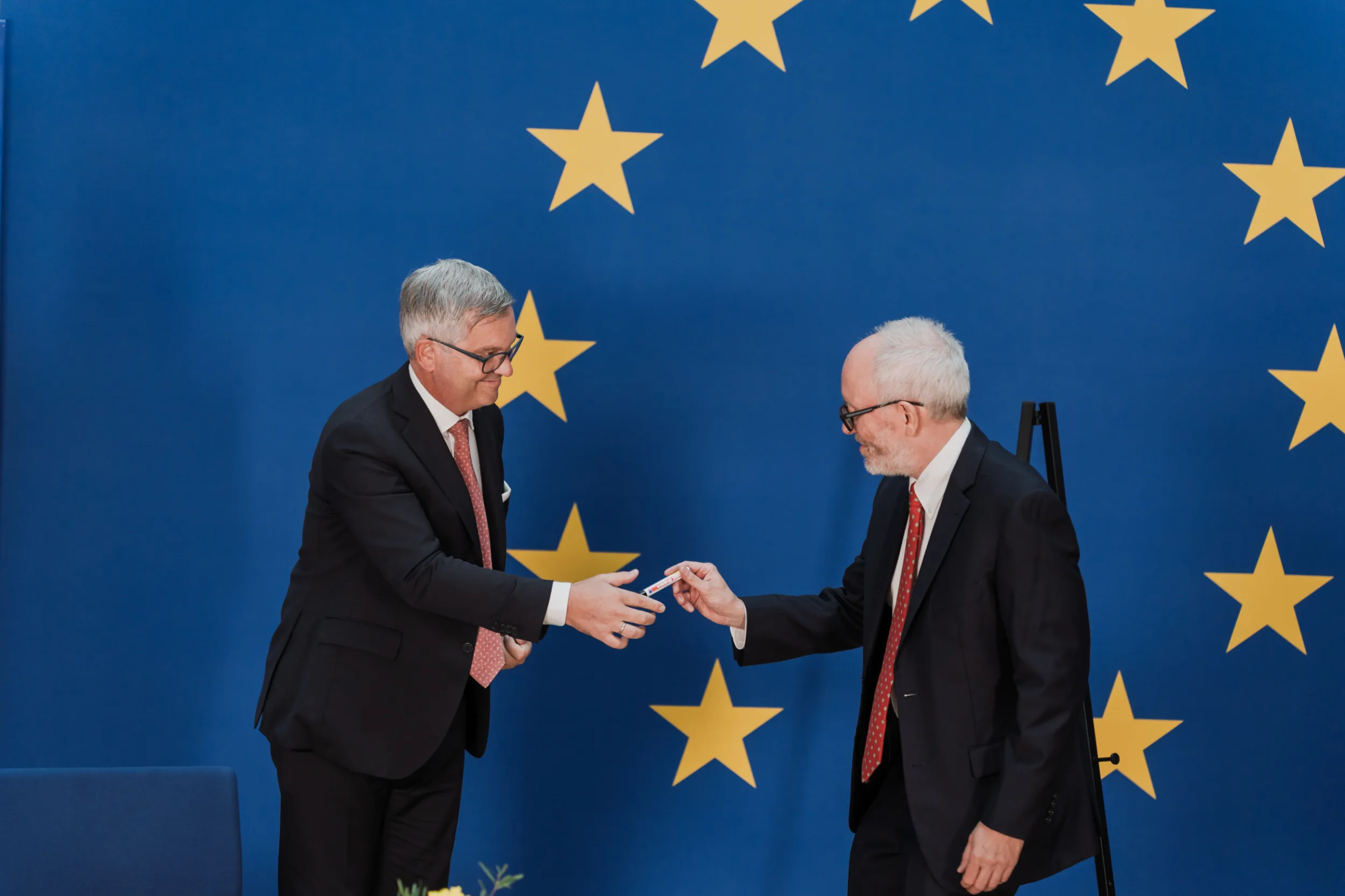 European Union Event Photographer | Grant Signing Ceremony in Vienna Fedor Vasilev Photographer In Berlin Man in a suit hands a pen to another man in a dark suit before a blue European Union star-themed backdrop.