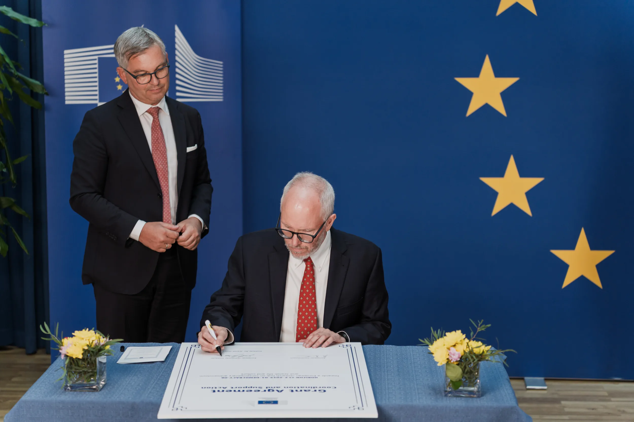 European Union Event Photographer | Grant Signing Ceremony in Vienna Fedor Vasilev Photographer In Berlin Man signs a Grant Agreement document at a table while another man in a suit watches before a blue EU star backdrop.