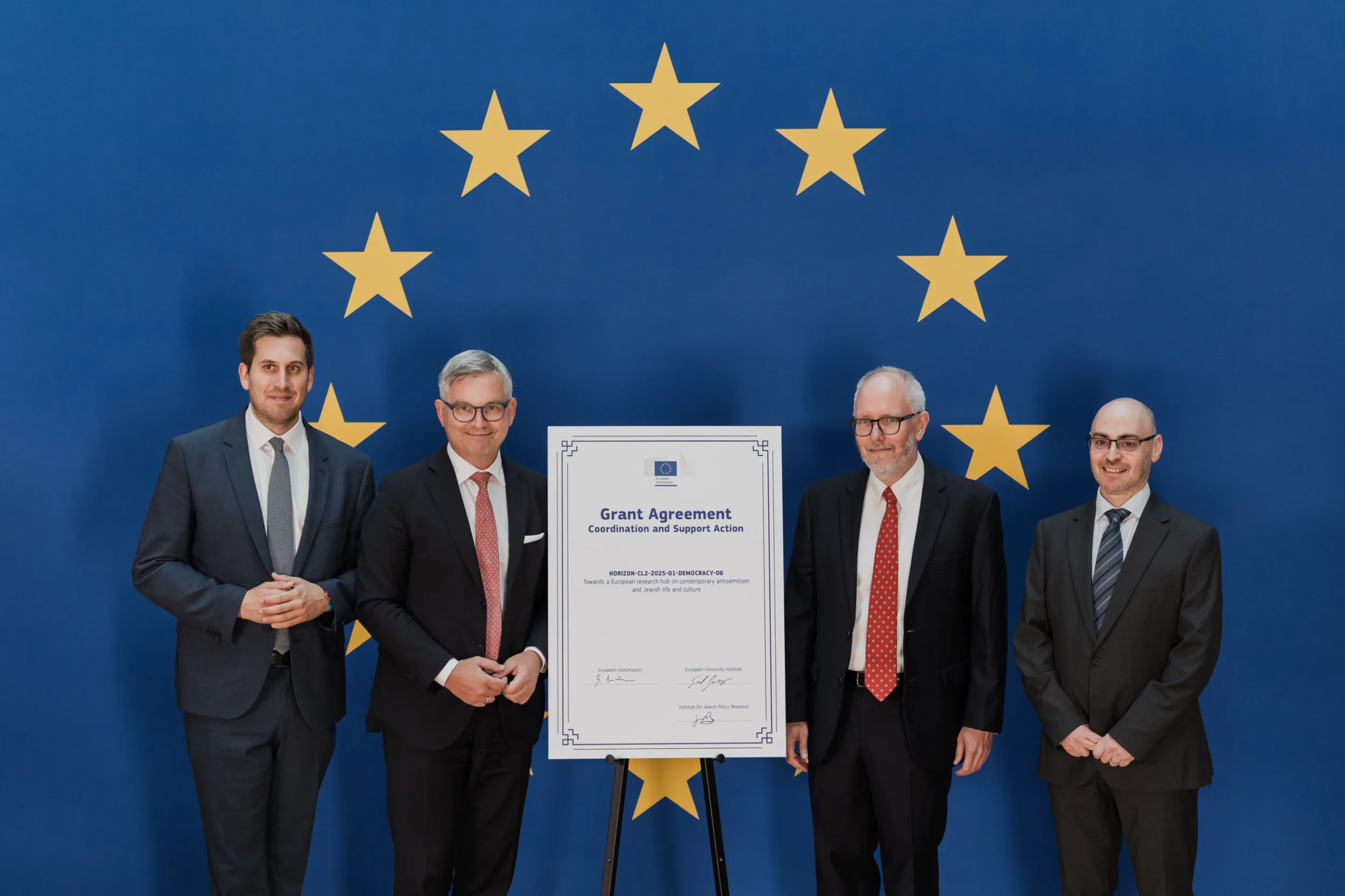 European Union Event Photographer | Grant Signing Ceremony in Vienna Fedor Vasilev Photographer In Berlin Four men in formal suits stand side-by-side behind a Grant Agreement document on an easel against a blue EU flag wall.
