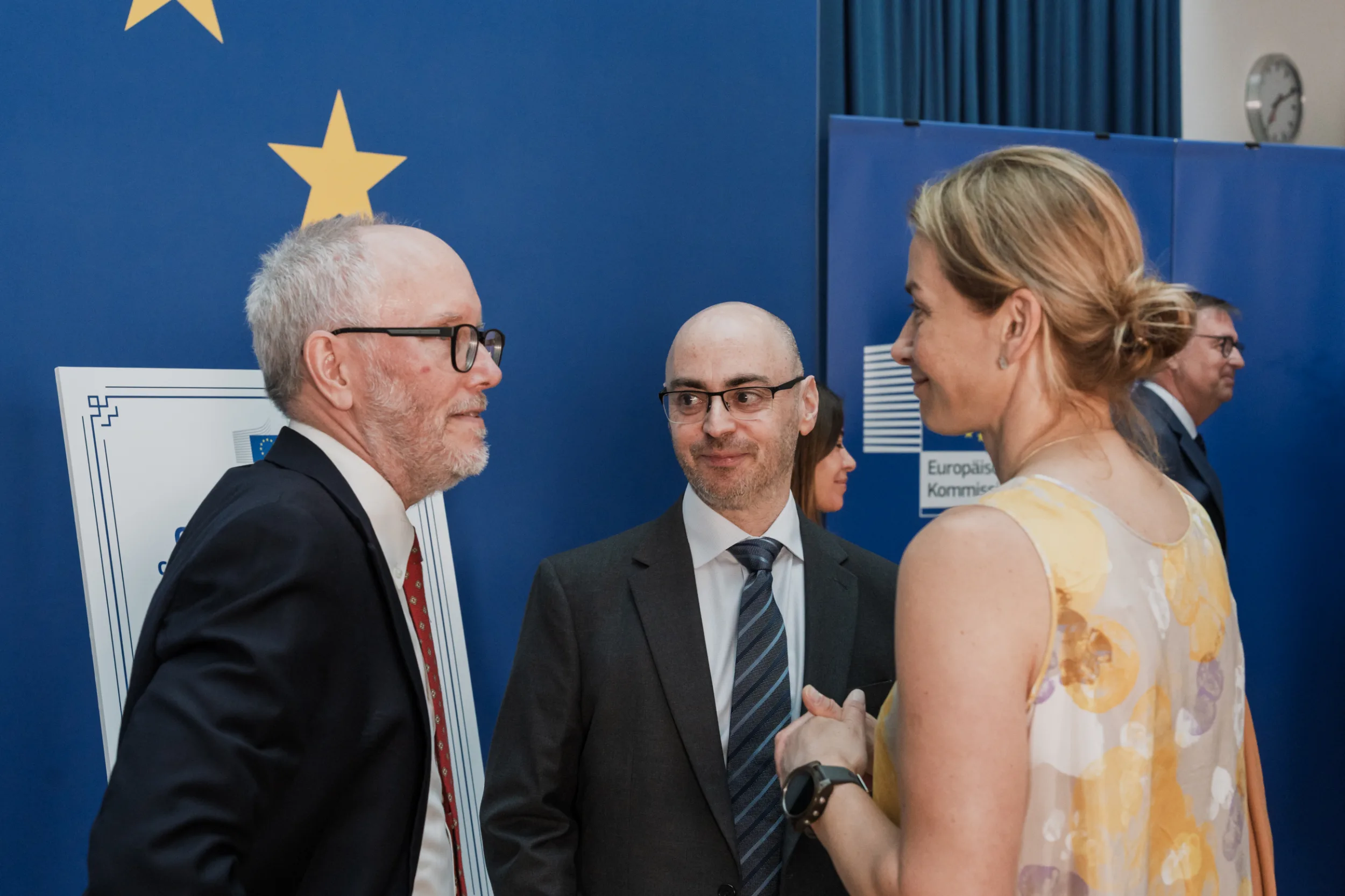 European Union Event Photographer | Grant Signing Ceremony in Vienna Fedor Vasilev Photographer In Berlin Two men in business suits and a woman in a floral top converse before a large blue European Union star-themed backdrop.
