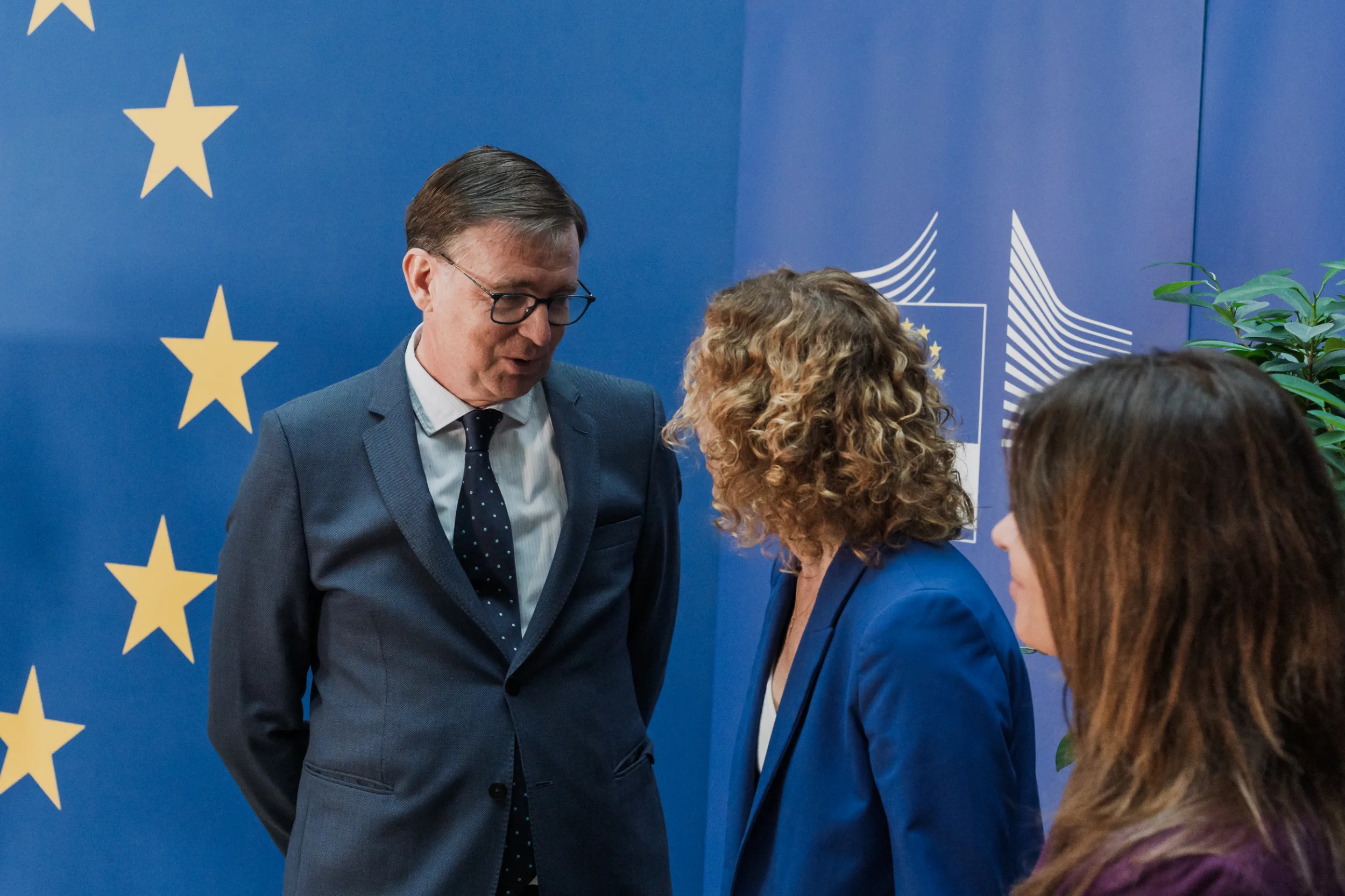 European Union Event Photographer | Grant Signing Ceremony in Vienna Fedor Vasilev Photographer In Berlin Man in a suit wearing glasses speaks to two women against a blue banner decorated with gold European Union stars.