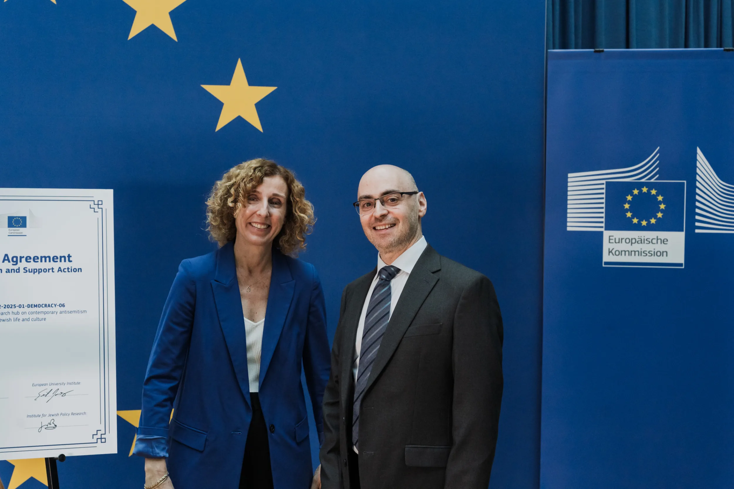 European Union Event Photographer | Grant Signing Ceremony in Vienna Fedor Vasilev Photographer In Berlin Woman in a blue blazer and man in a suit pose before a European Commission banner and a grant agreement document.