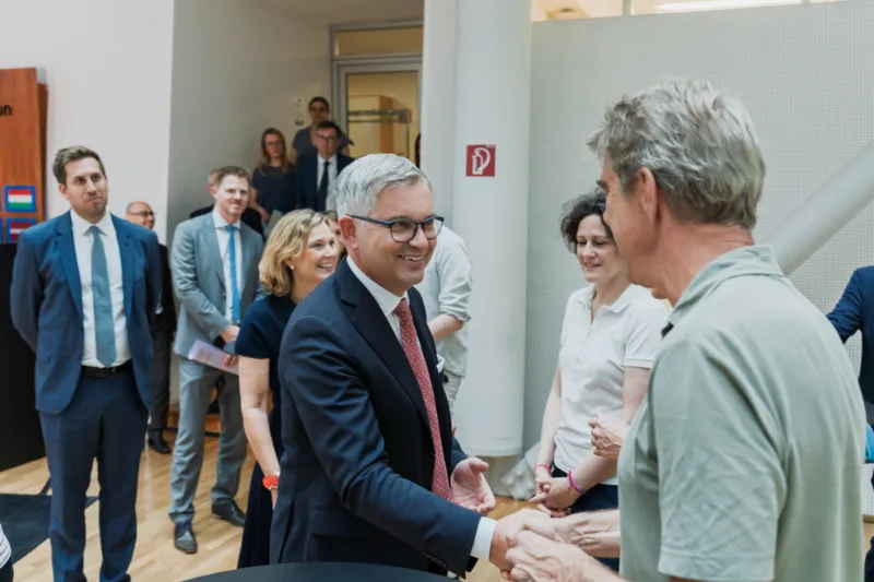 Man in a suit and glasses shakes hands with another person at a formal indoor event while others observe.