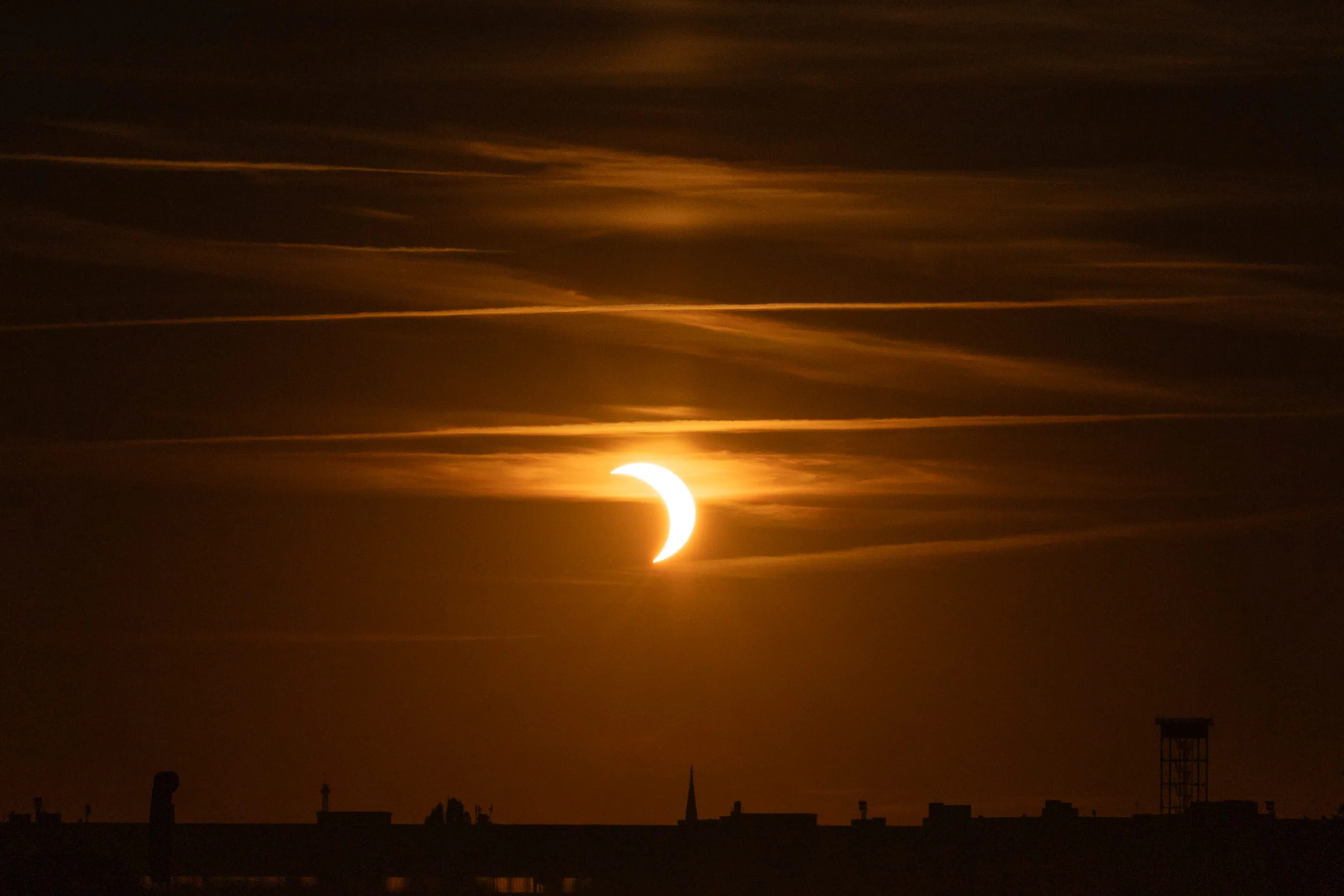 Photographing the Solar Eclipse in Berlin Fedor Vasilev Photographer In Berlin Crescent sun glows through dark, horizontal cloud layers above a distant, jagged city skyline silhouette.