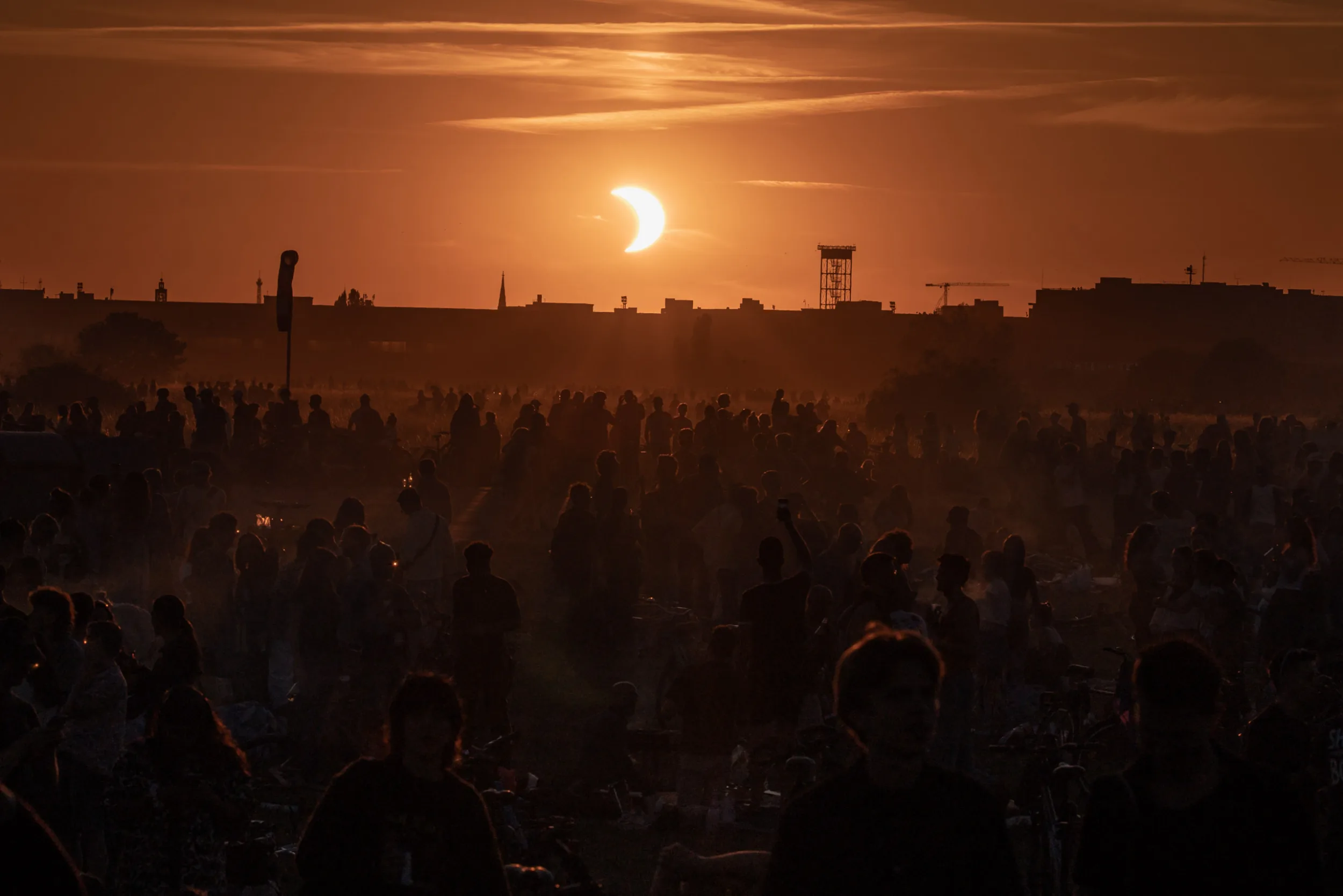 Crowd of people silhouetted against a golden sunset during a partial solar eclipse over the Berlin city skyline. Photographed from Tempelhofer Feld