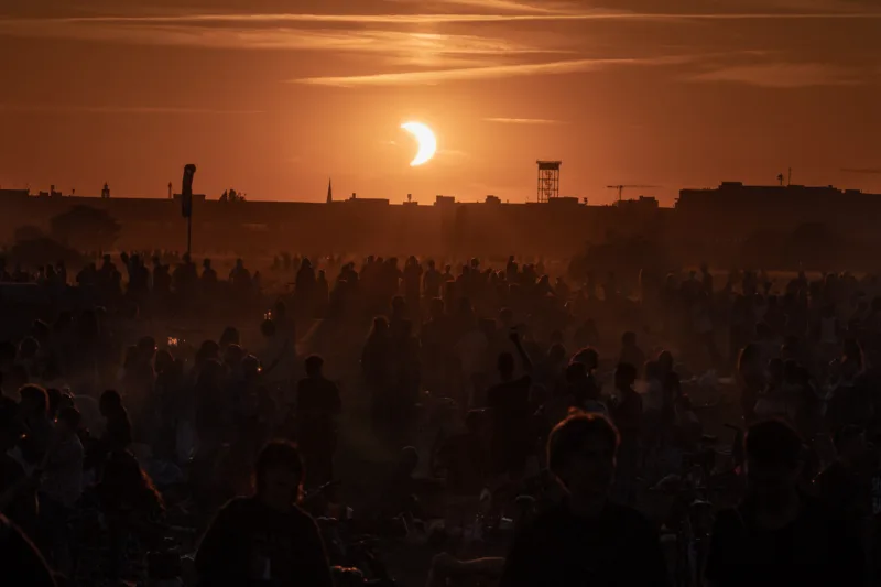 Crowd of people silhouetted against a golden sunset during a partial solar eclipse over the Berlin city skyline. Photographed from Tempelhofer Feld