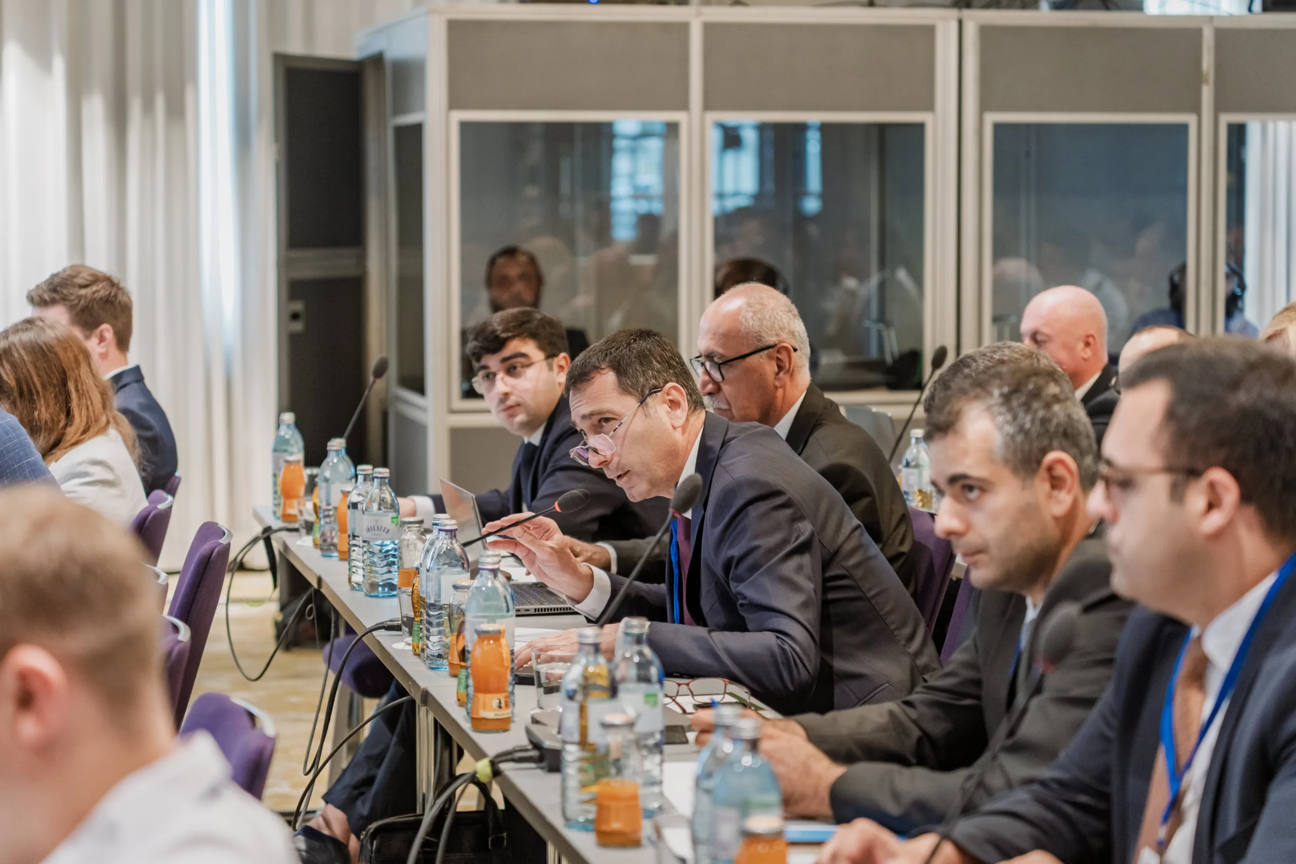 EU4Energy Conference Photography in Vienna Fedor Vasilev Photographer In Berlin Professional in a suit speaks into a conference microphone while sitting at a long table with colleagues.