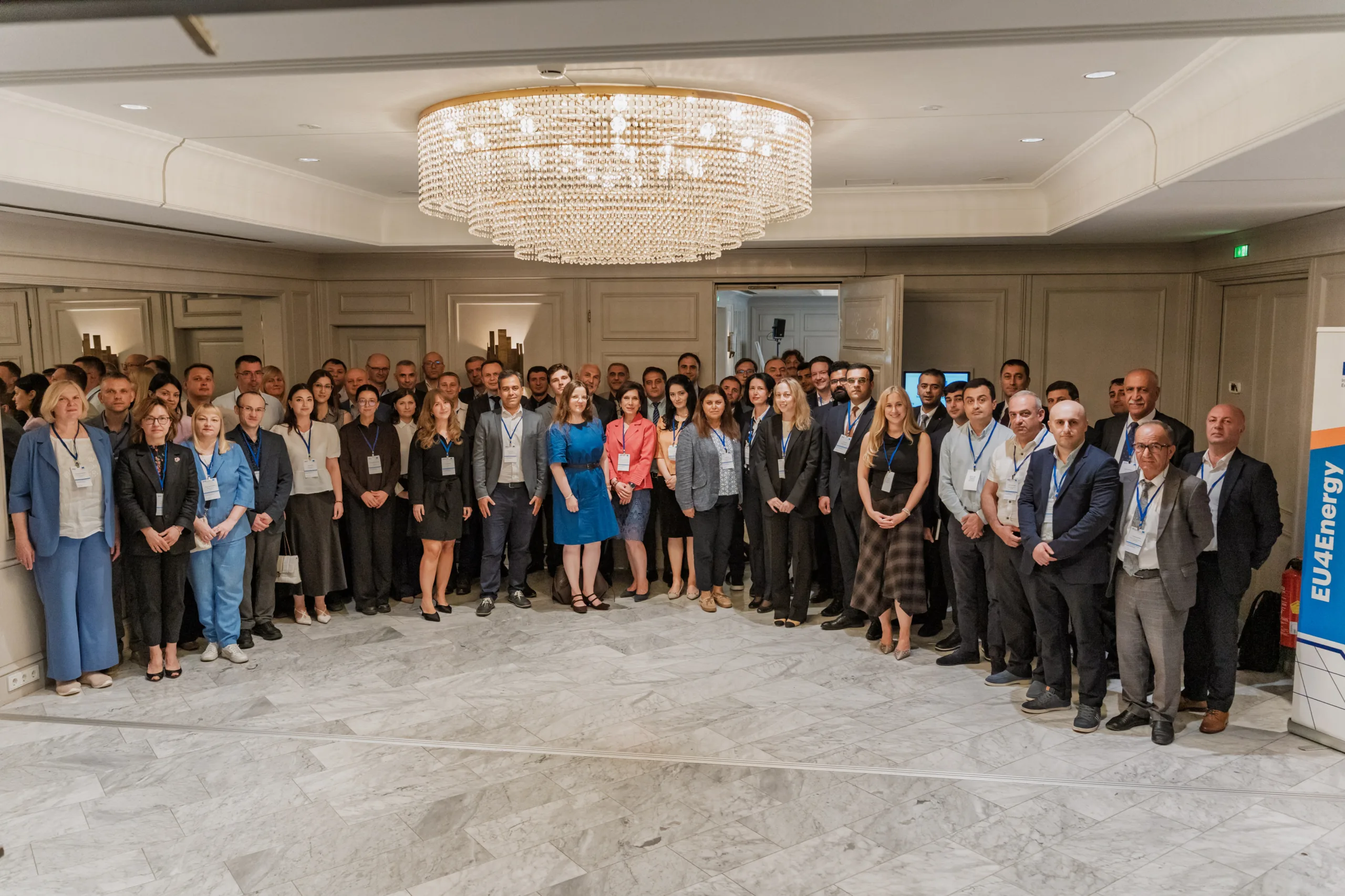 EU4Energy Conference Photography in Vienna Fedor Vasilev Photographer In Berlin Large group of conference attendees posing on marble flooring beneath a crystal chandelier near an EU4Energy banner.