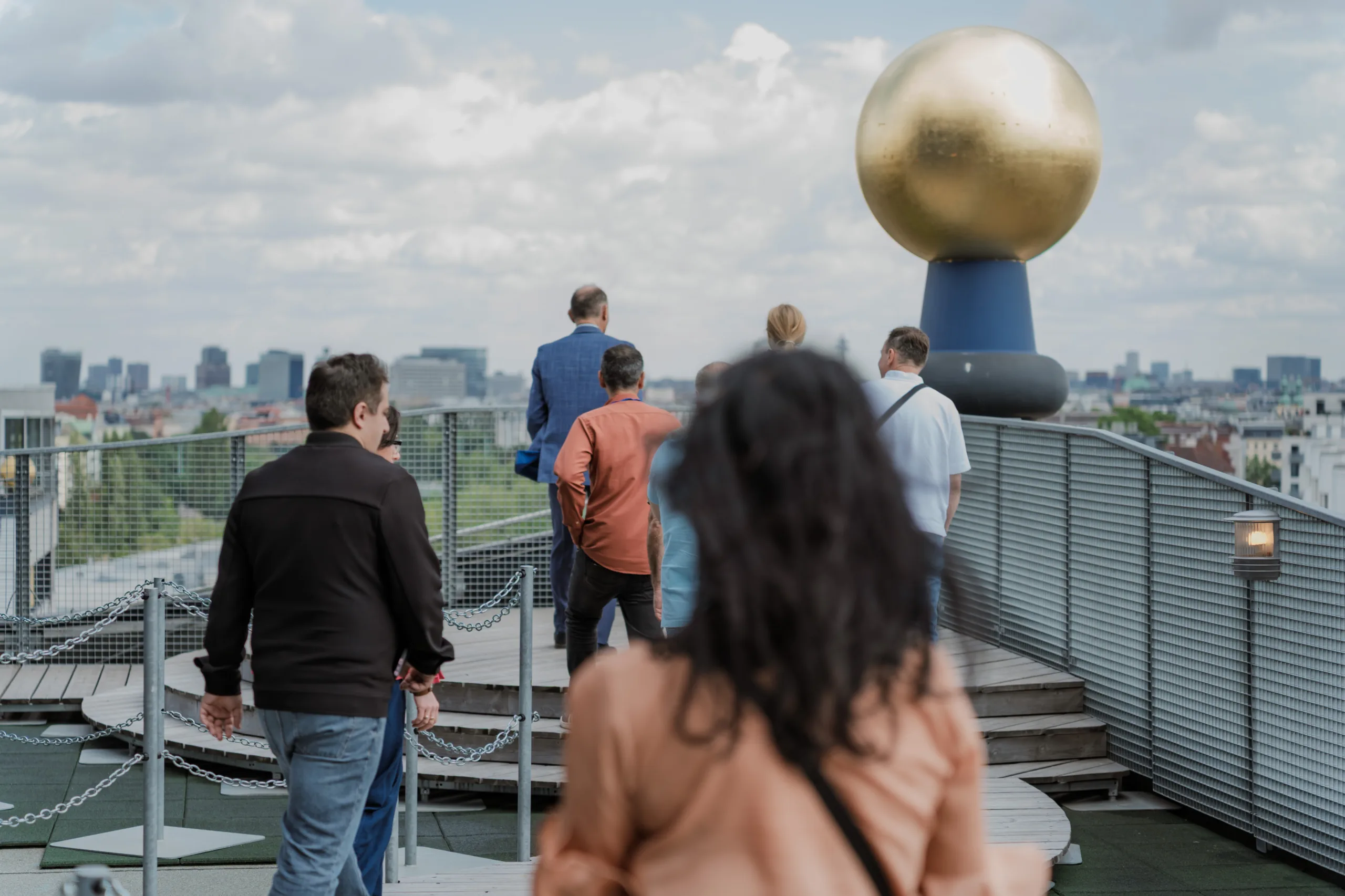 EU4Energy Conference Photography in Vienna Fedor Vasilev Photographer In Berlin People walk across a rooftop terrace towards a large golden spherical structure overlooking the city skyline.