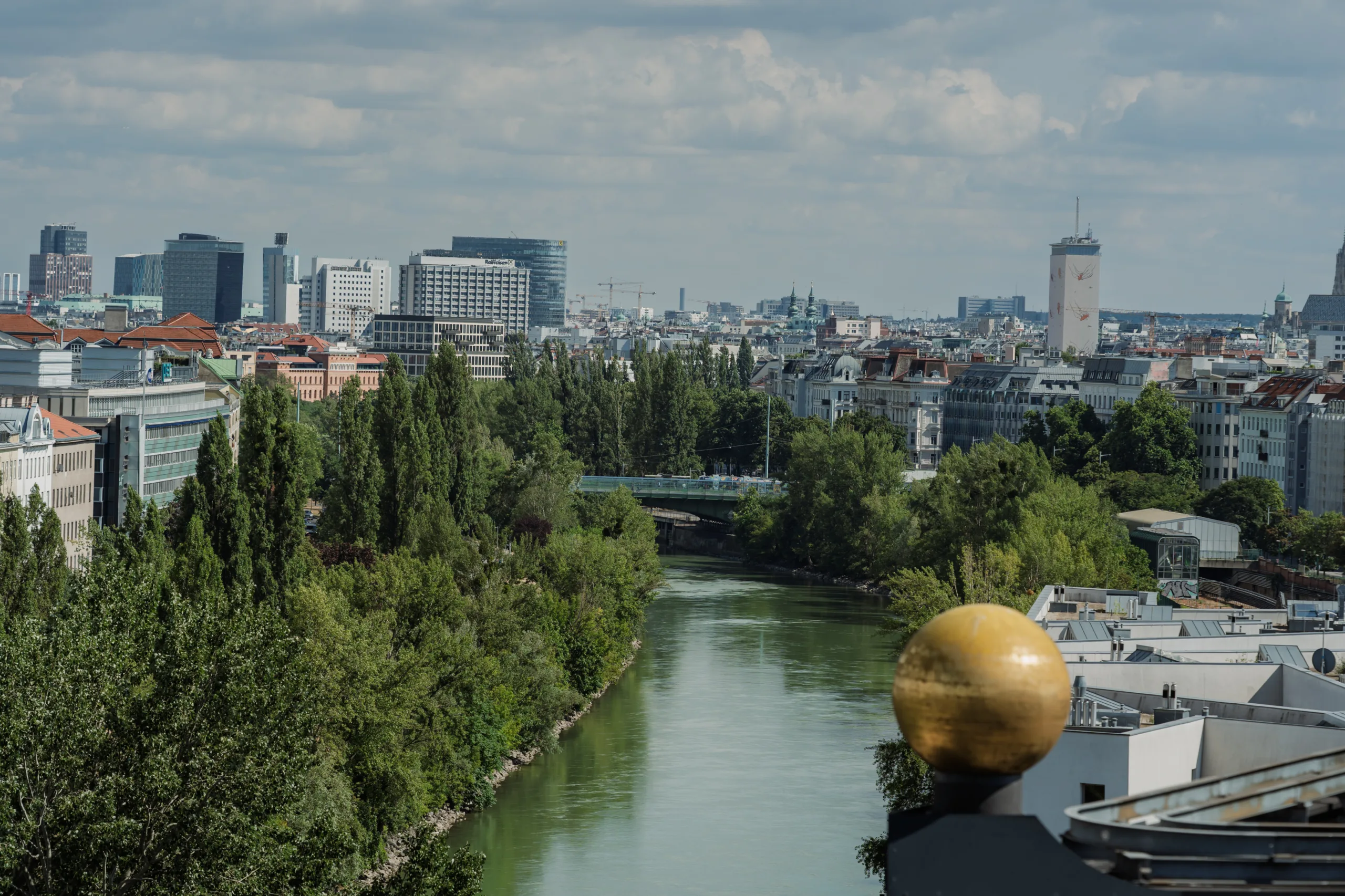 EU4Energy Conference Photography in Vienna Fedor Vasilev Photographer In Berlin Panoramic view of a city river flowing past dense green trees and urban buildings under a cloudy sky.
