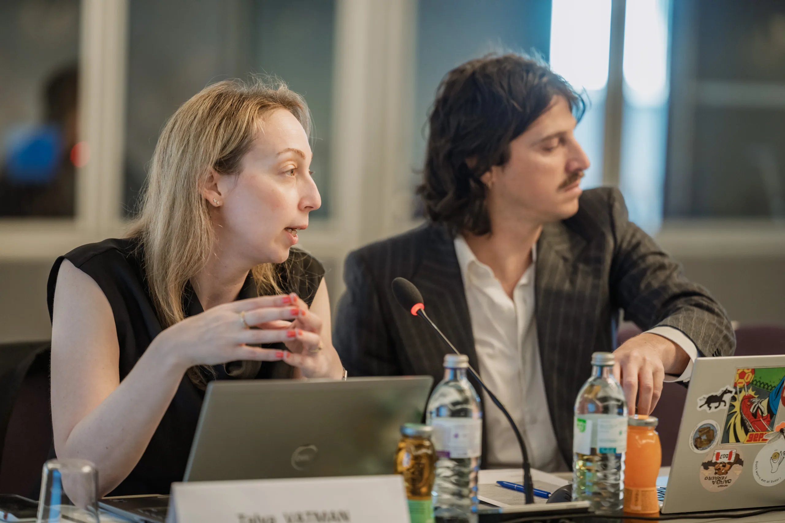 EU4Energy Conference Photography in Vienna Fedor Vasilev Photographer In Berlin Blonde woman in a black top gestures while speaking next to a man with long hair sitting at a table with laptops.