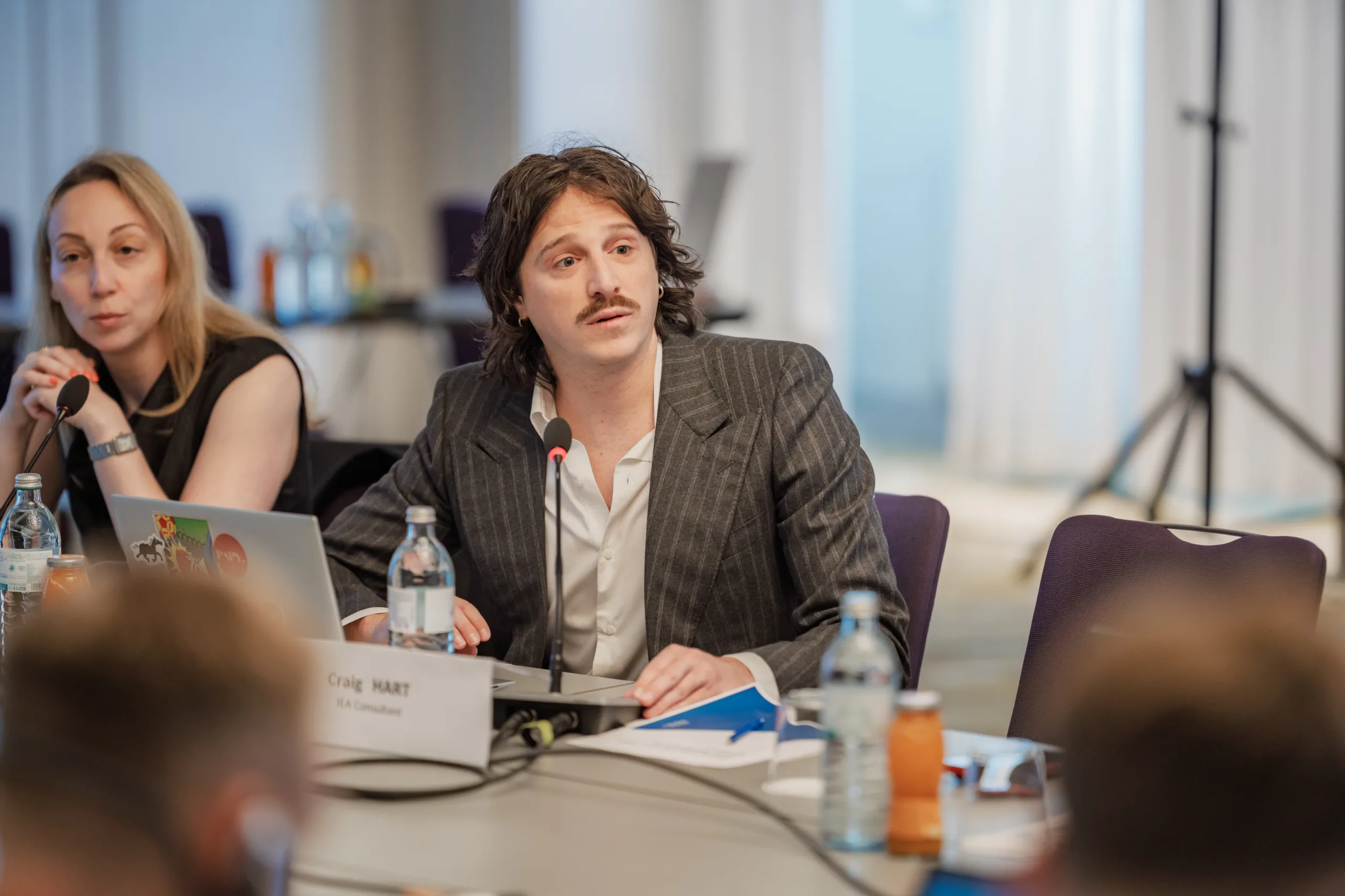 EU4Energy Conference Photography in Vienna Fedor Vasilev Photographer In Berlin Long-haired man with a mustache in a striped blazer sits at a conference table with a name card reading Craig Hart.