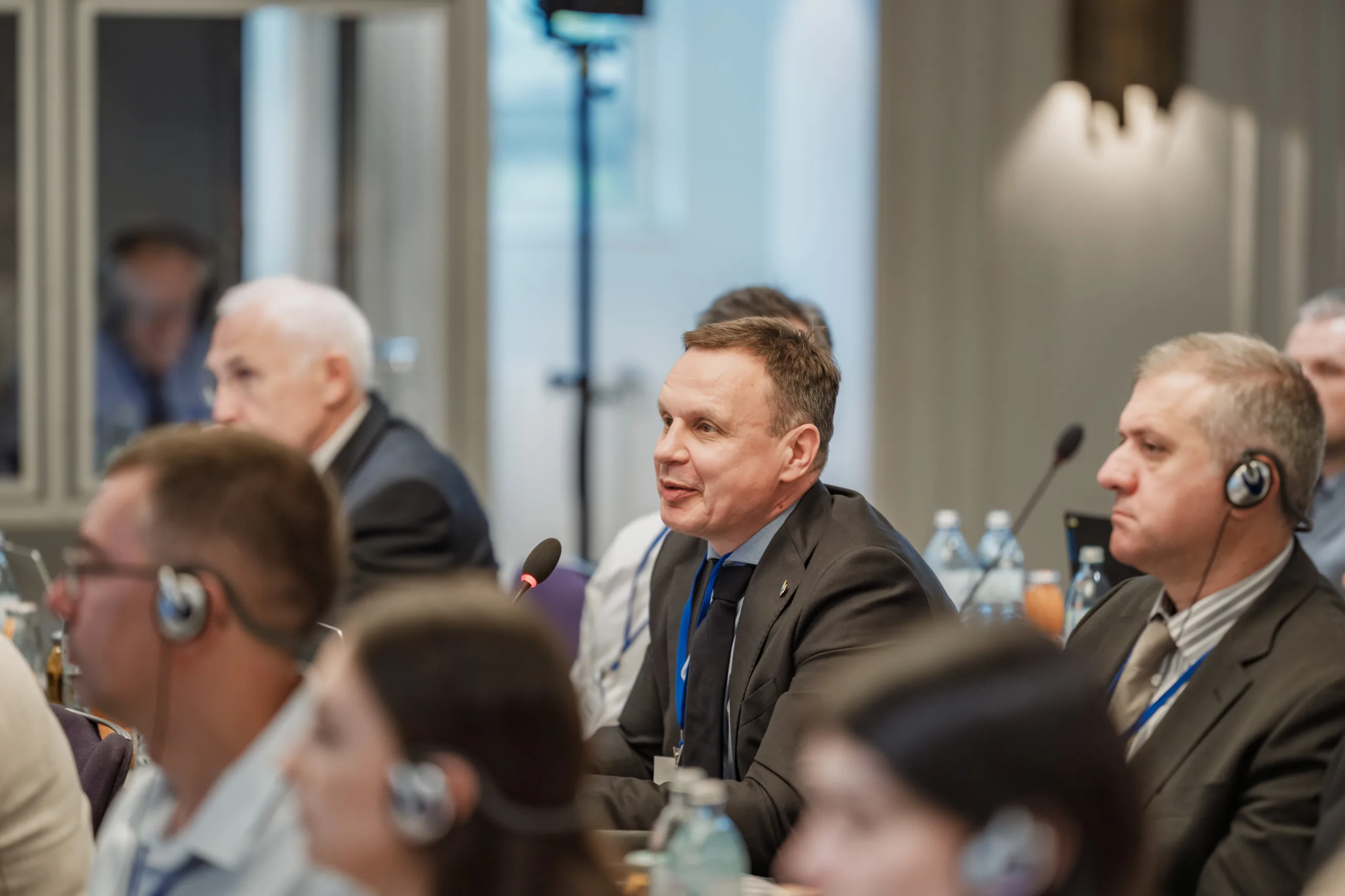 EU4Energy Conference Photography in Vienna Fedor Vasilev Photographer In Berlin Attendee wearing a dark suit and tie shares thoughts into a microphone while others listen at a conference table.