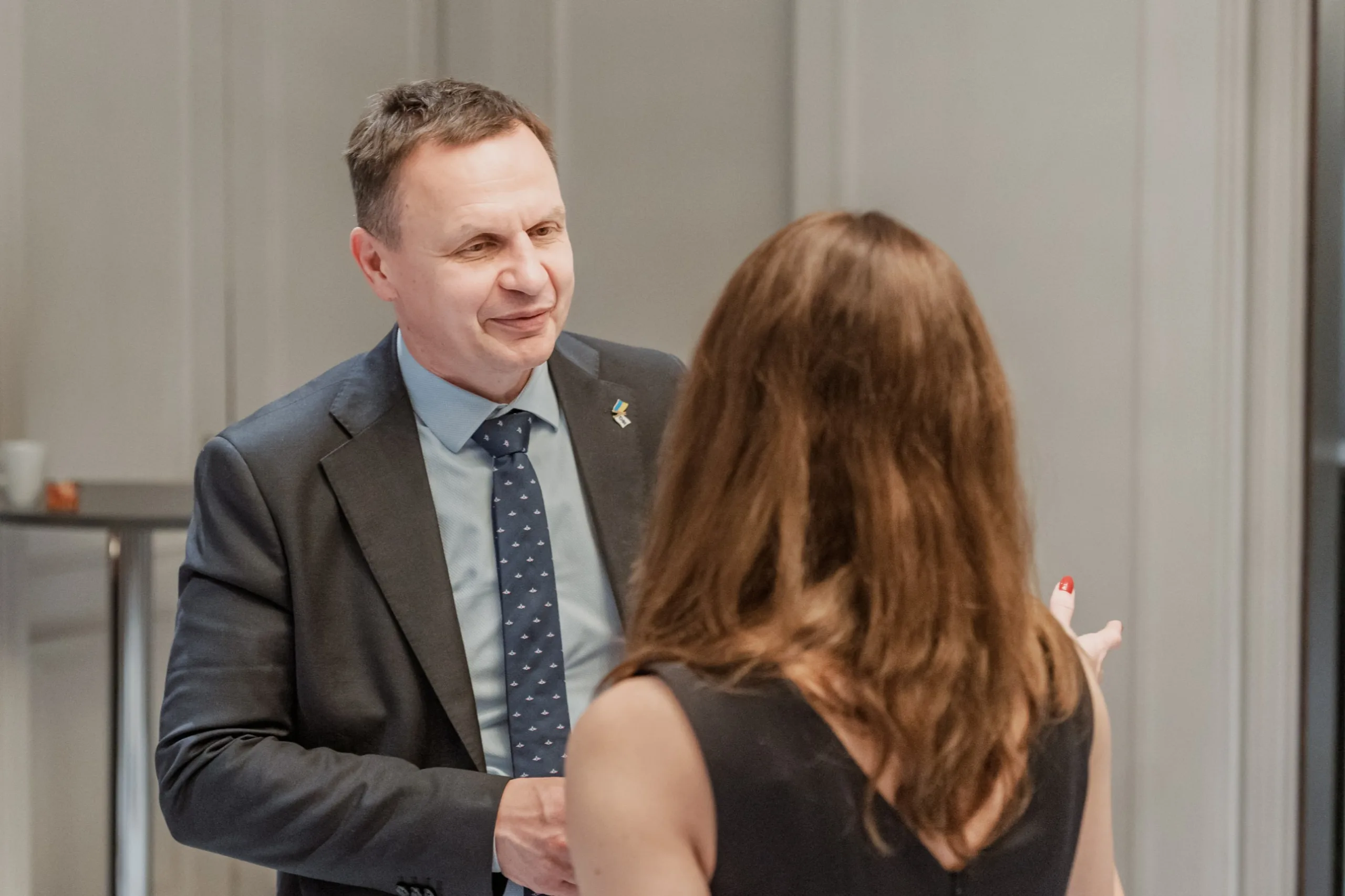 EU4Energy Conference Photography in Vienna Fedor Vasilev Photographer In Berlin Smiling man in a dark suit and patterned tie gestures while speaking to a woman in a black sleeveless dress.