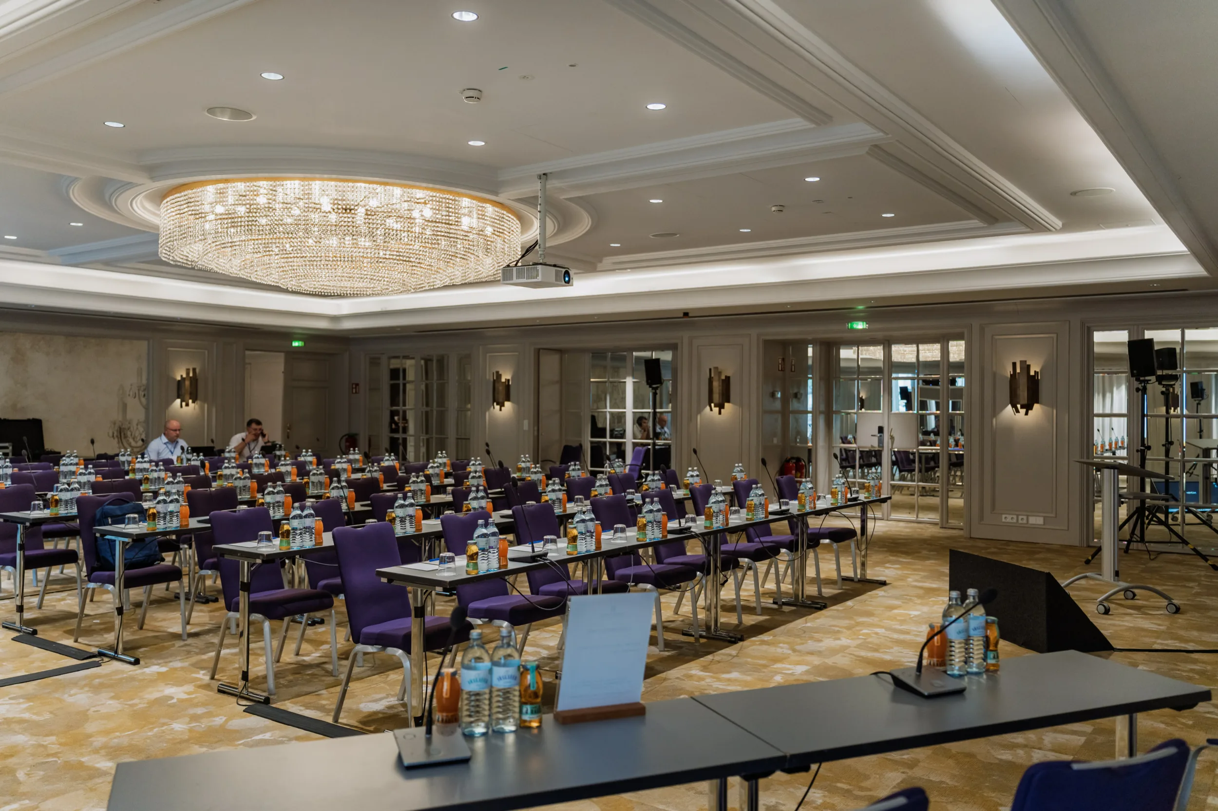 Rows of purple chairs and tables set up for a corporate meeting under a large crystal chandelier in a hotel ballroom.