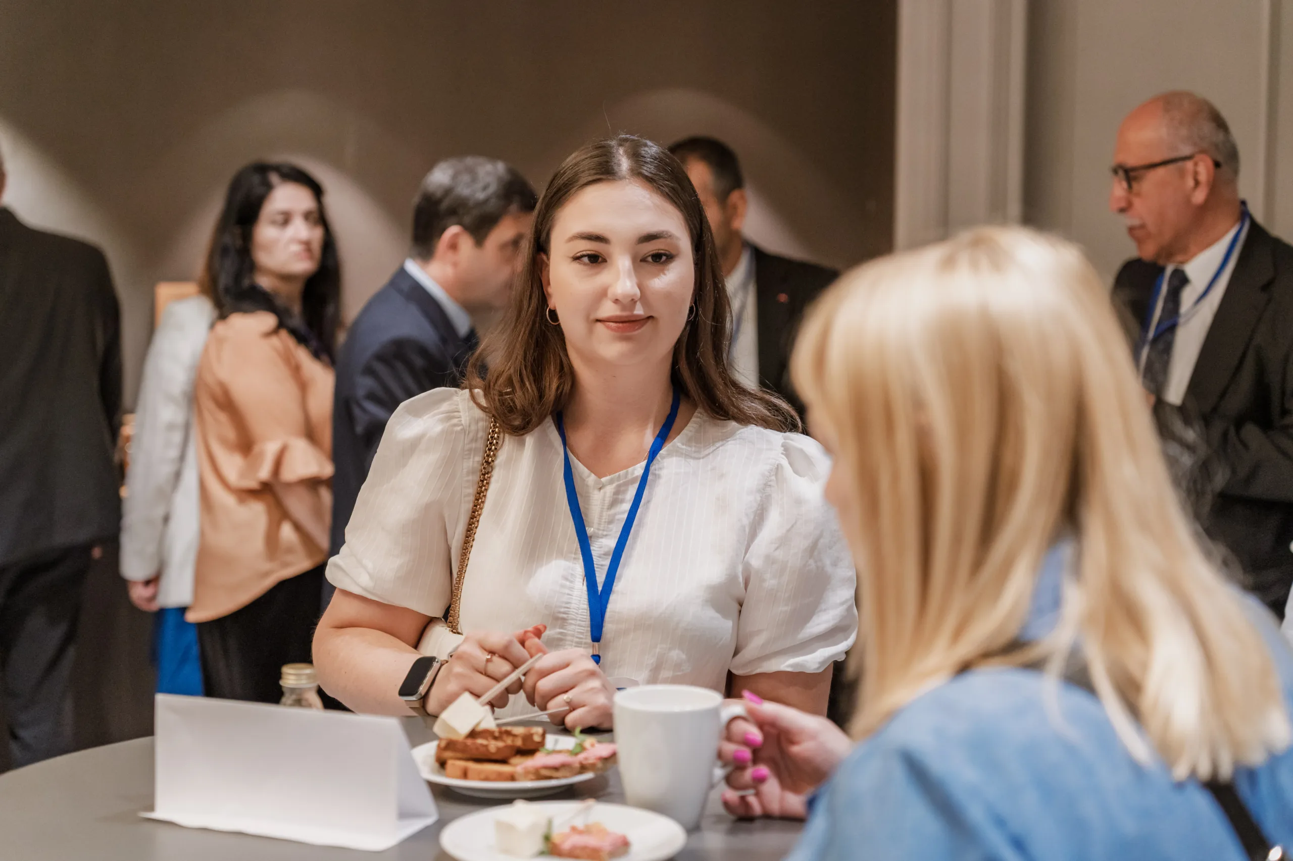 EU4Energy Conference Photography in Vienna Fedor Vasilev Photographer In Berlin Young woman in a white blouse with a blue lanyard smiles while enjoying a snack and coffee during a conference break.