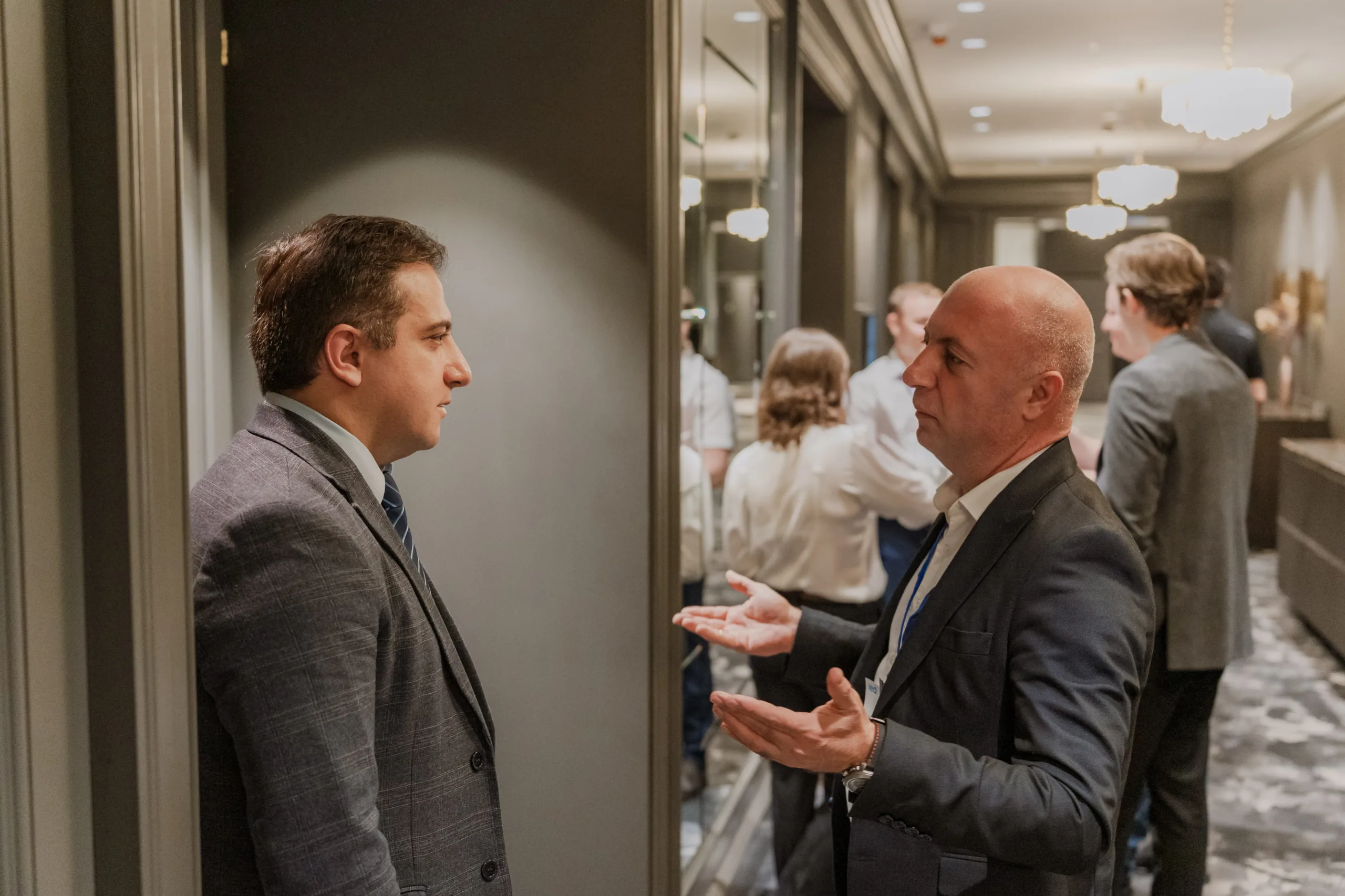 EU4Energy Conference Photography in Vienna Fedor Vasilev Photographer In Berlin Two men in dark suits engage in a focused conversation near a hallway entrance while other attendees mingle in the background.