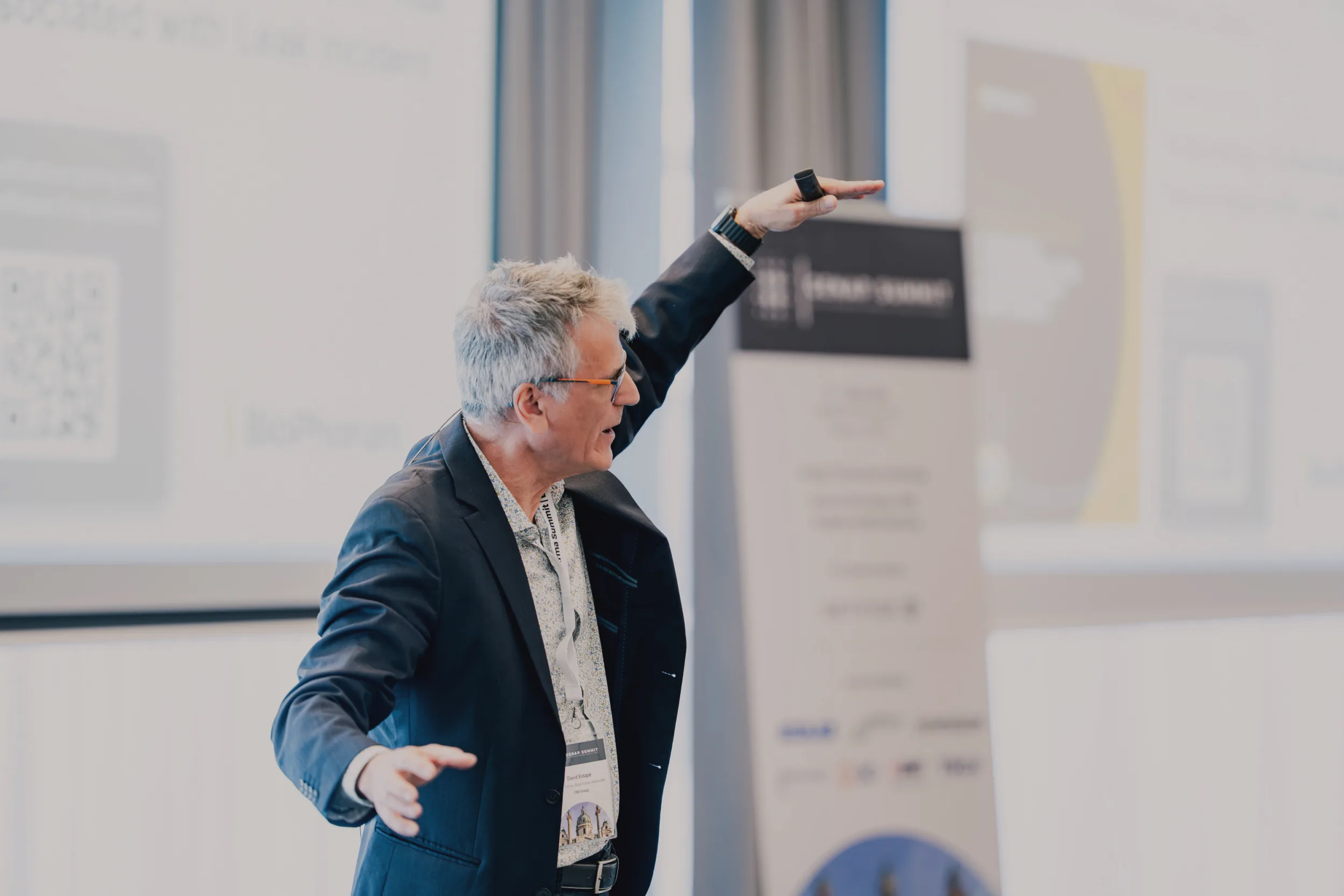 Genup Summit | Medical Conference Photography and Product Expo Fedor Vasilev Photographer In Berlin Silver-haired man in a dark blazer gestures upward while presenting at a Genup Summit event.