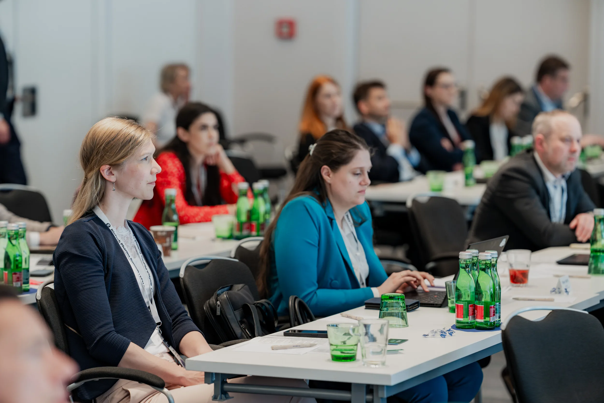 Genup Summit | Medical Conference Photography and Product Expo Fedor Vasilev Photographer In Berlin Attendees seated at tables observe a presentation while a woman in a blue blazer types on her laptop.