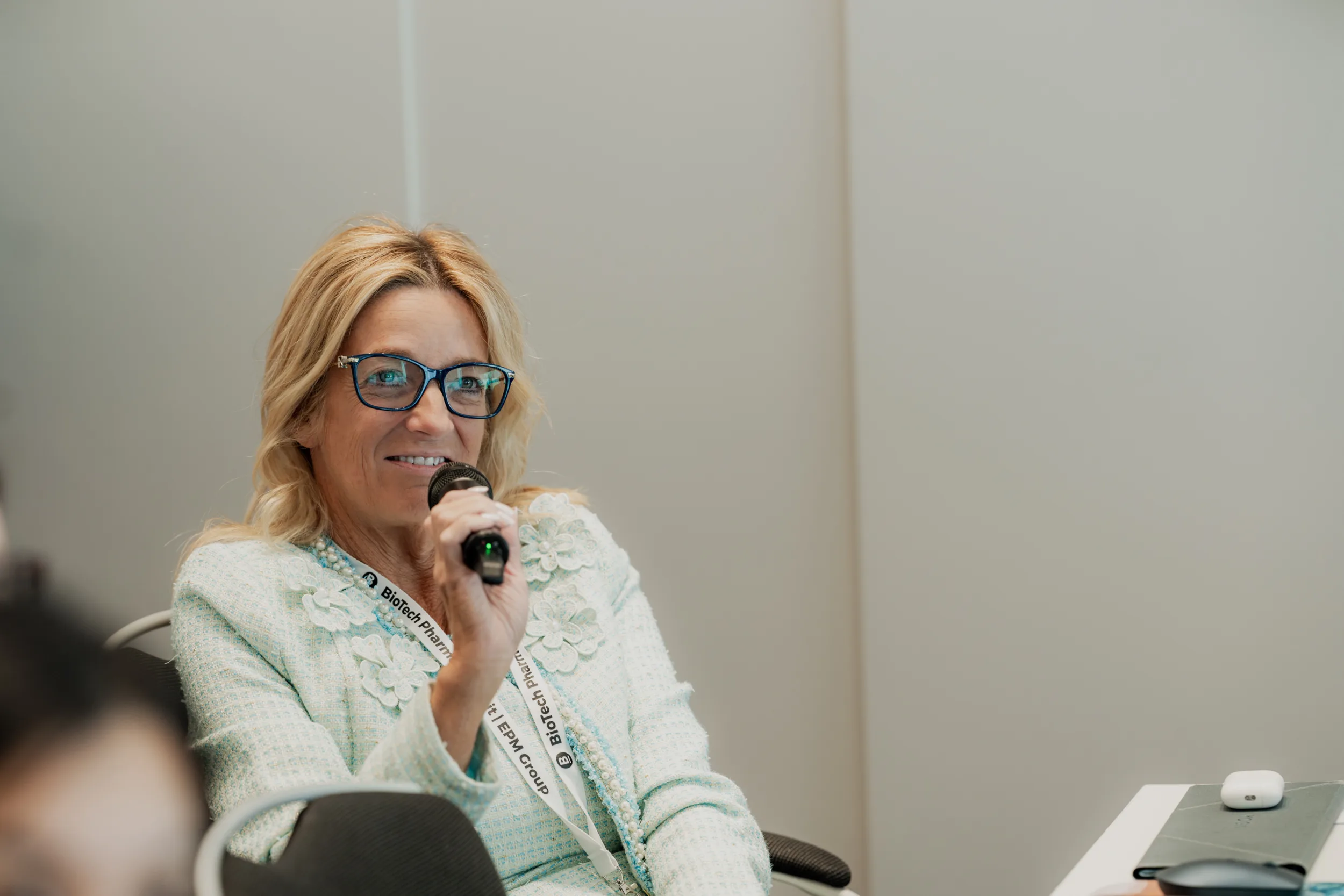 Genup Summit | Medical Conference Photography and Product Expo Fedor Vasilev Photographer In Berlin Woman wearing glasses and a light blue floral-detailed blazer speaks into a microphone at a professional event.