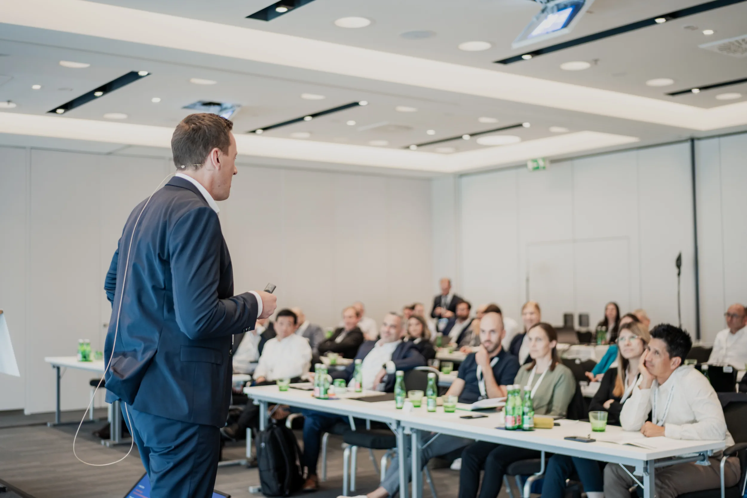 Genup Summit | Medical Conference Photography and Product Expo Fedor Vasilev Photographer In Berlin Man in a navy blazer wearing a headset microphone addresses an attentive audience seated at tables in a bright conference.