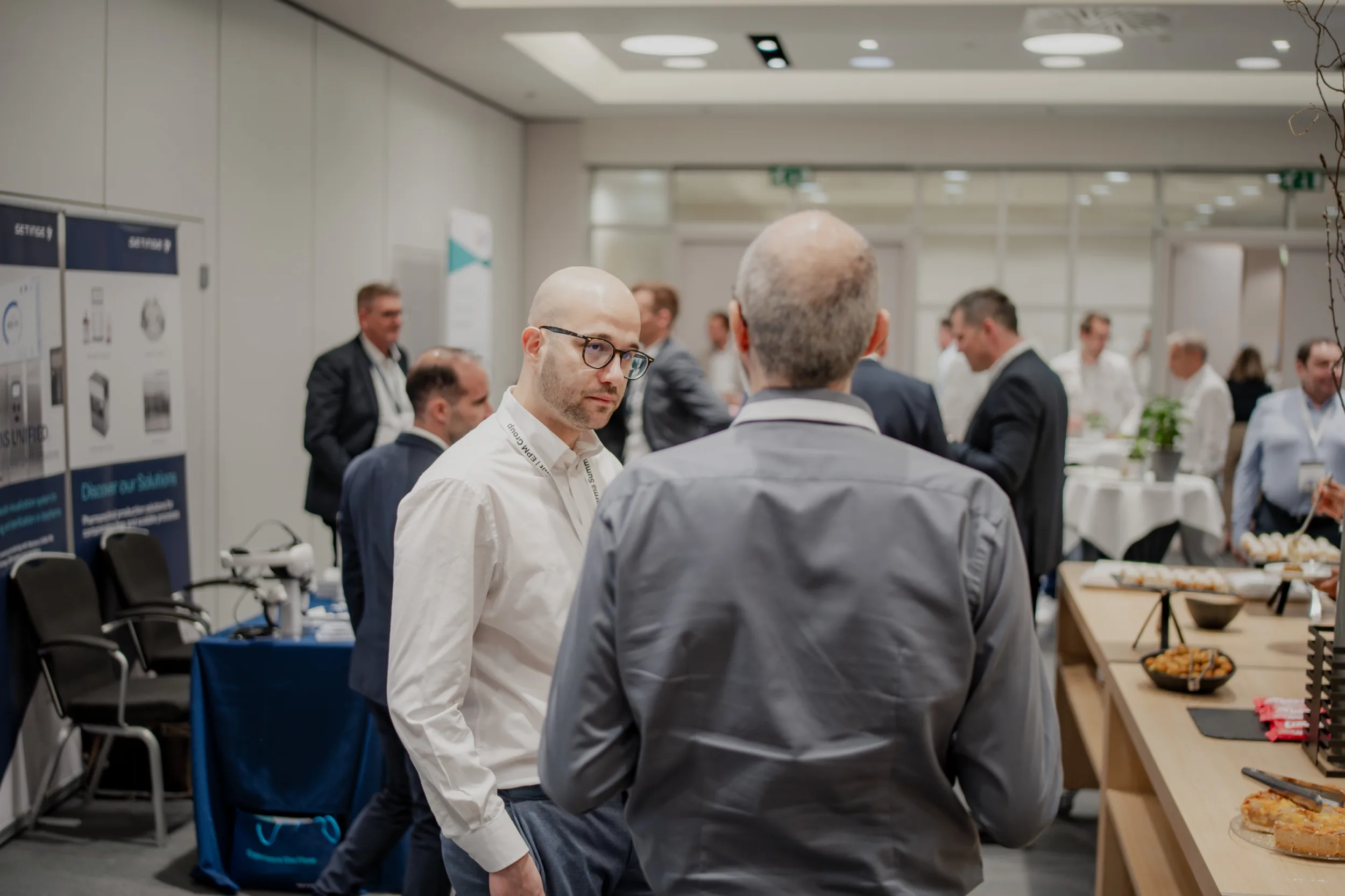 Genup Summit | Medical Conference Photography and Product Expo Fedor Vasilev Photographer In Berlin Bald man in glasses and white shirt listens to a colleague near a catering table at a busy conference.