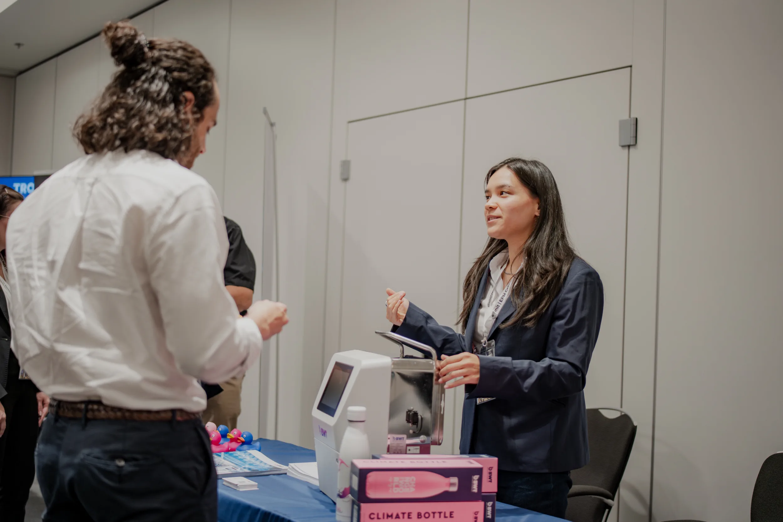 Genup Summit | Medical Conference Photography and Product Expo Fedor Vasilev Photographer In Berlin Young woman in a dark blazer explains a medical device to a man with long hair tied in a bun at an expo booth.
