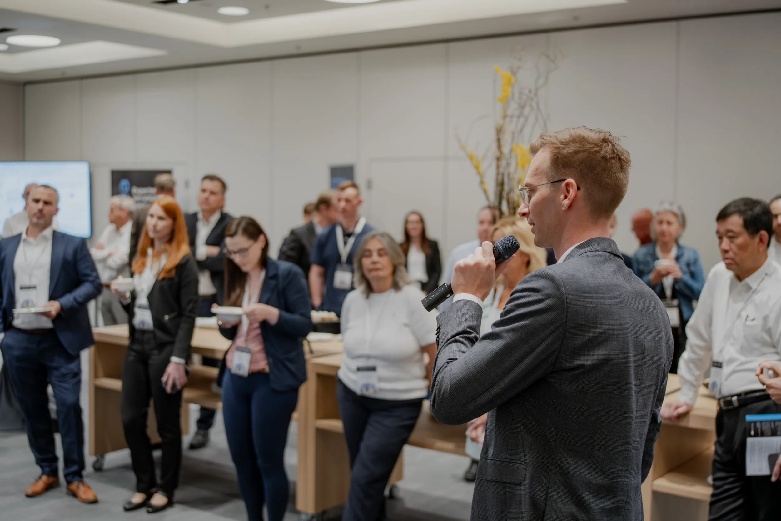 Genup Summit | Medical Conference Photography and Product Expo Fedor Vasilev Photographer In Berlin Man in a grey blazer speaks into a microphone while facing a group of attendees at a conference gathering.