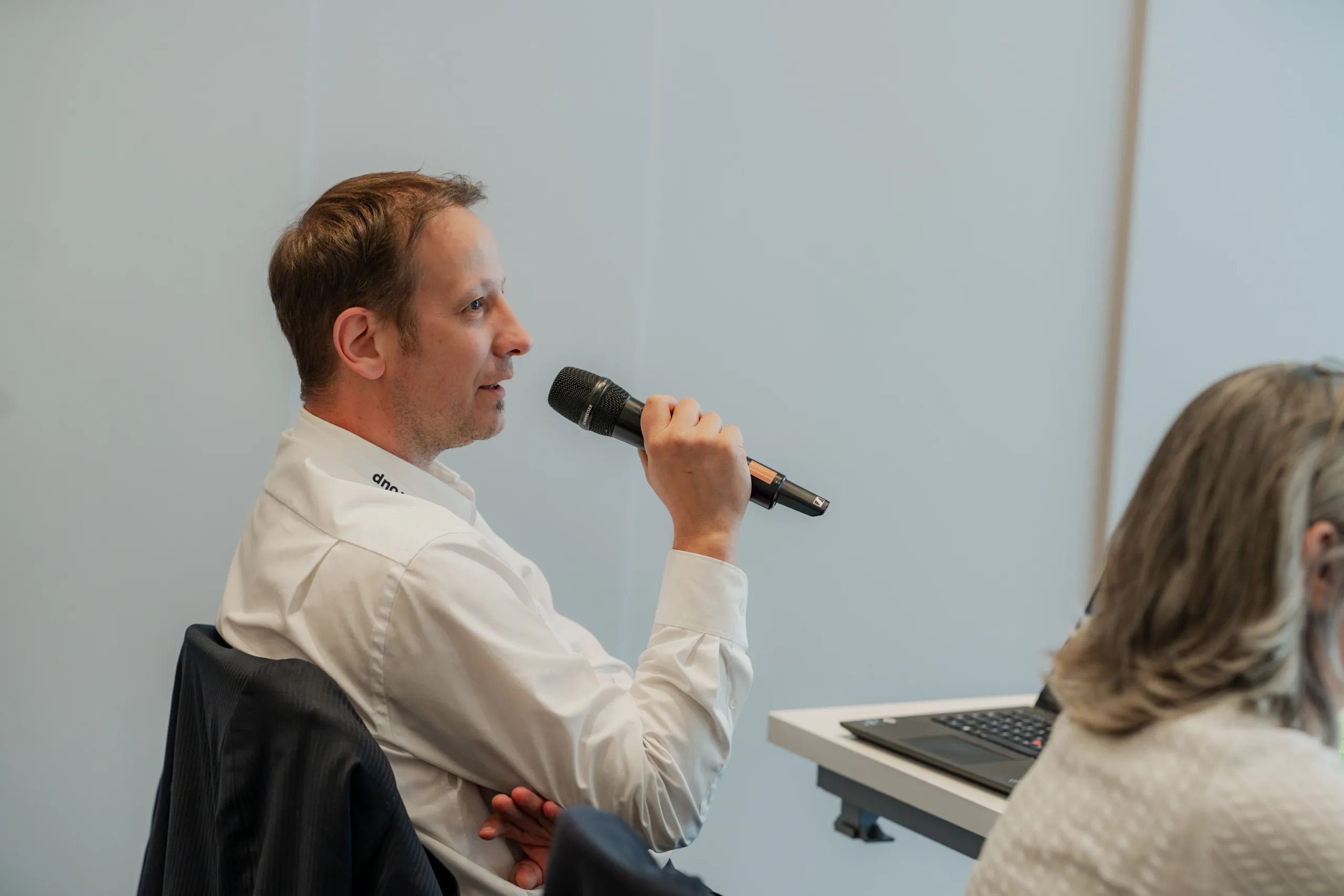 Genup Summit | Medical Conference Photography and Product Expo Fedor Vasilev Photographer In Berlin Man in a white shirt holds a microphone to speak while seated at a table during a meeting.