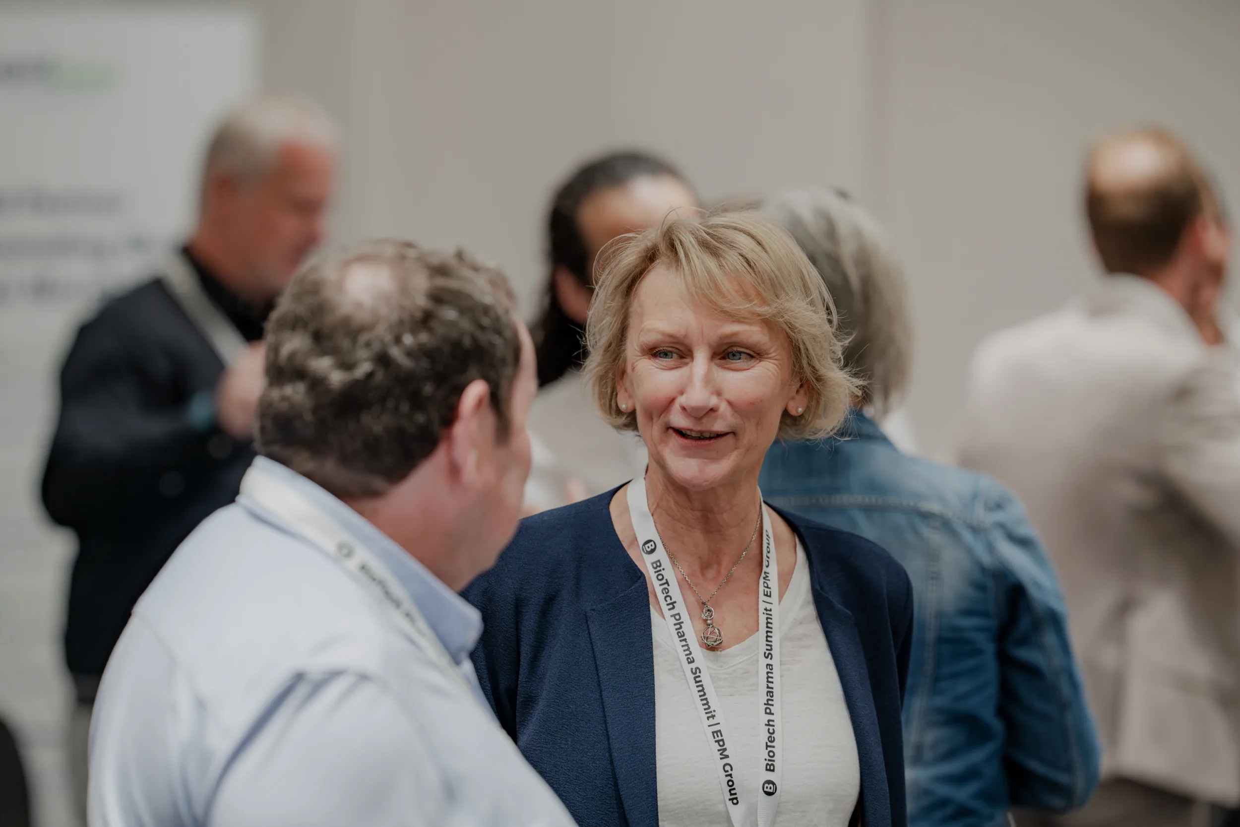 Genup Summit | Medical Conference Photography and Product Expo Fedor Vasilev Photographer In Berlin Blonde woman wearing a BioTech Pharma Summit lanyard smiles while chatting with a man at a professional gathering.