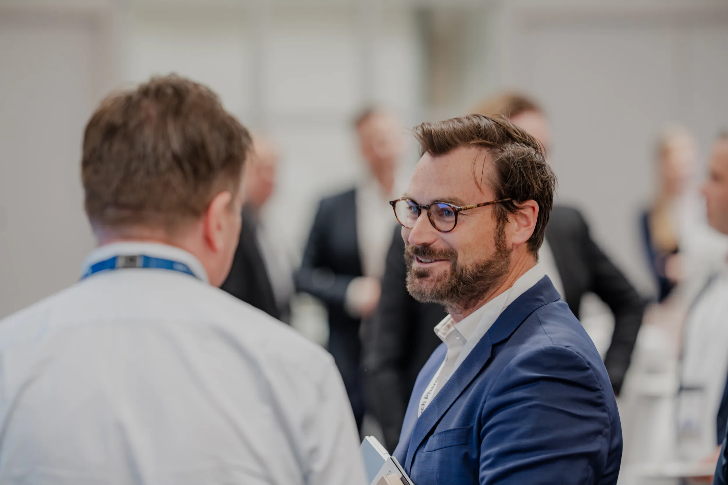 Genup Summit | Medical Conference Photography and Product Expo Fedor Vasilev Photographer In Berlin Bearded man in a navy blazer and tortoise-shell glasses smiles while talking to a colleague at the Genup Summit.