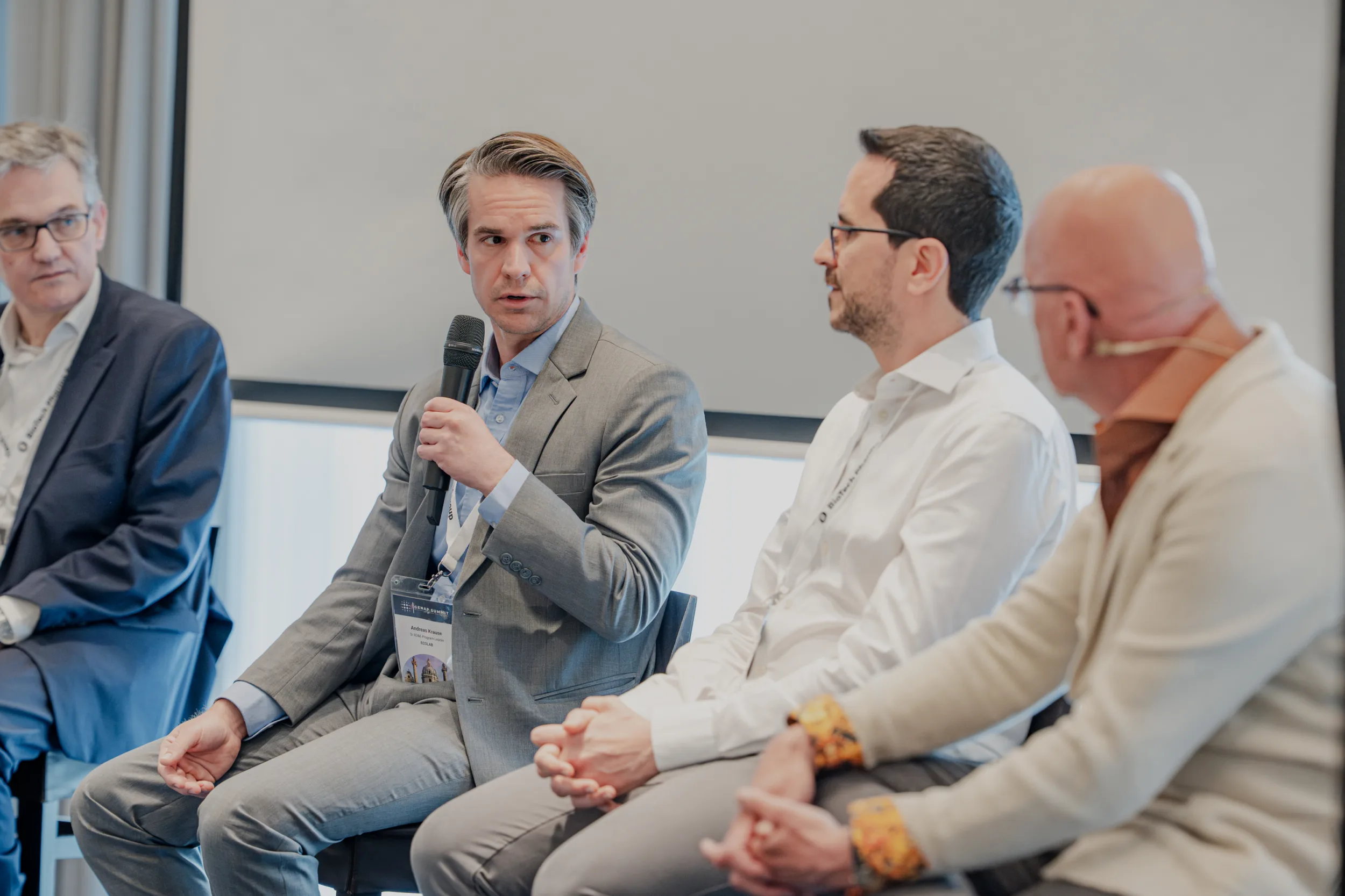 Genup Summit | Medical Conference Photography and Product Expo Fedor Vasilev Photographer In Berlin Panelists in business attire sit on a stage during a conference while one man speaks into a handheld microphone.