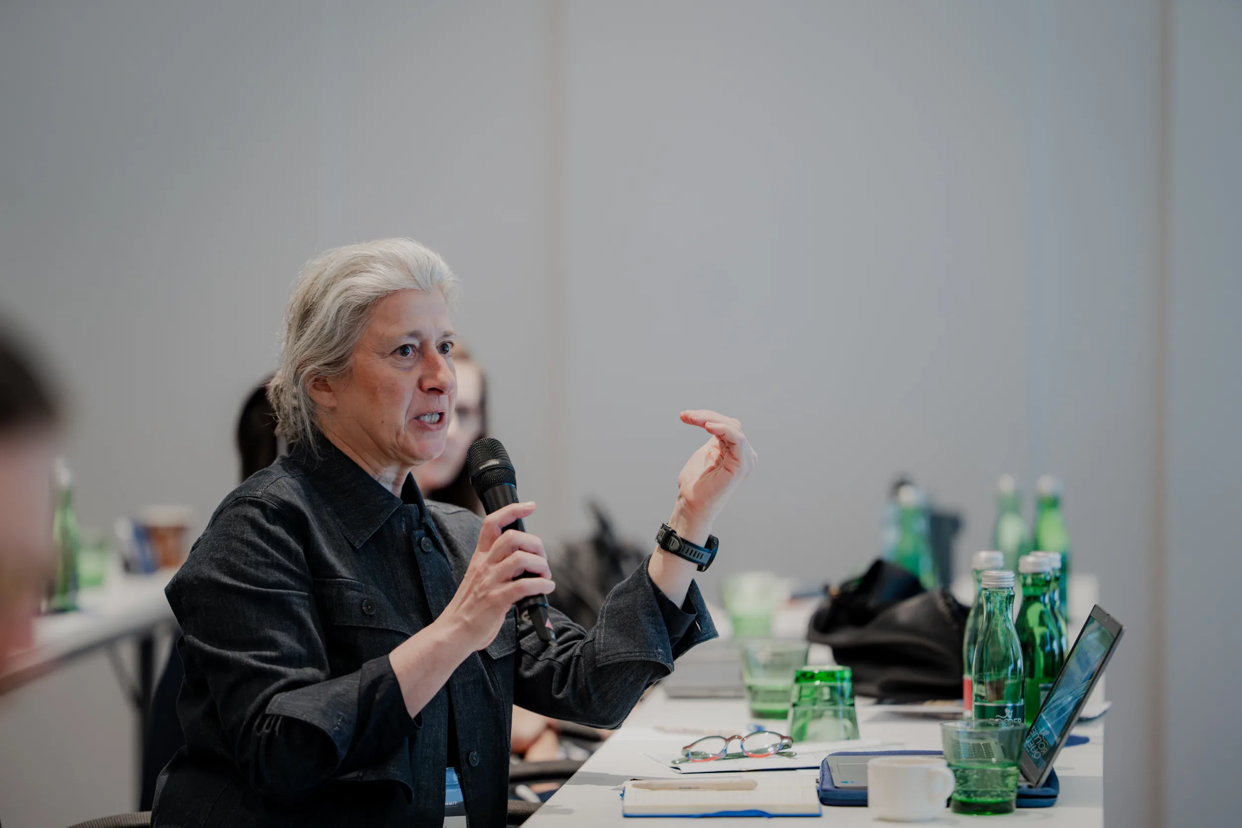 Genup Summit | Medical Conference Photography and Product Expo Fedor Vasilev Photographer In Berlin Silver-haired woman in a black jacket gestures while speaking into a handheld microphone at a meeting table.