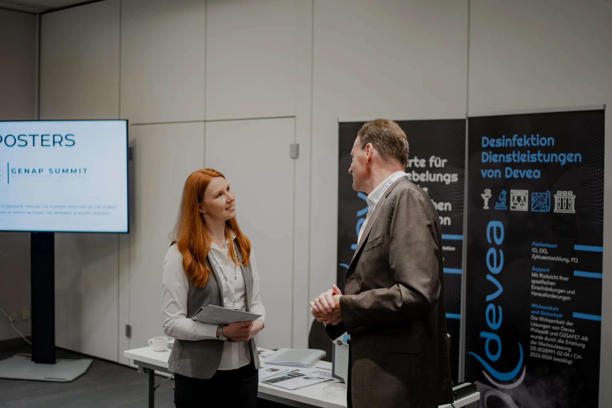 Genup Summit | Medical Conference Photography and Product Expo Fedor Vasilev Photographer In Berlin Red-haired woman in a grey vest talks to a man in a brown suit beside a Devea branded banner at a conference.
